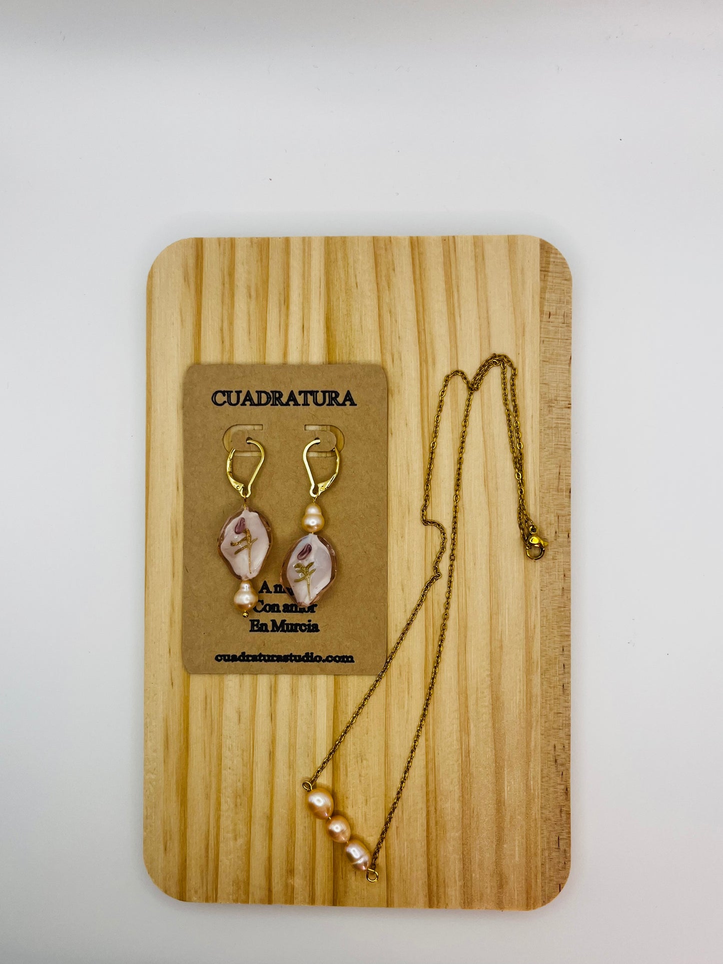 Conjunto artesanal Bella: colgante con tres perlas melocotón en cadena dorada y pendientes de cristal blanco con motivo floral y perla melocotón, en tarjeta Cuadratura sobre tabla de madera, montados a mano en Murcia