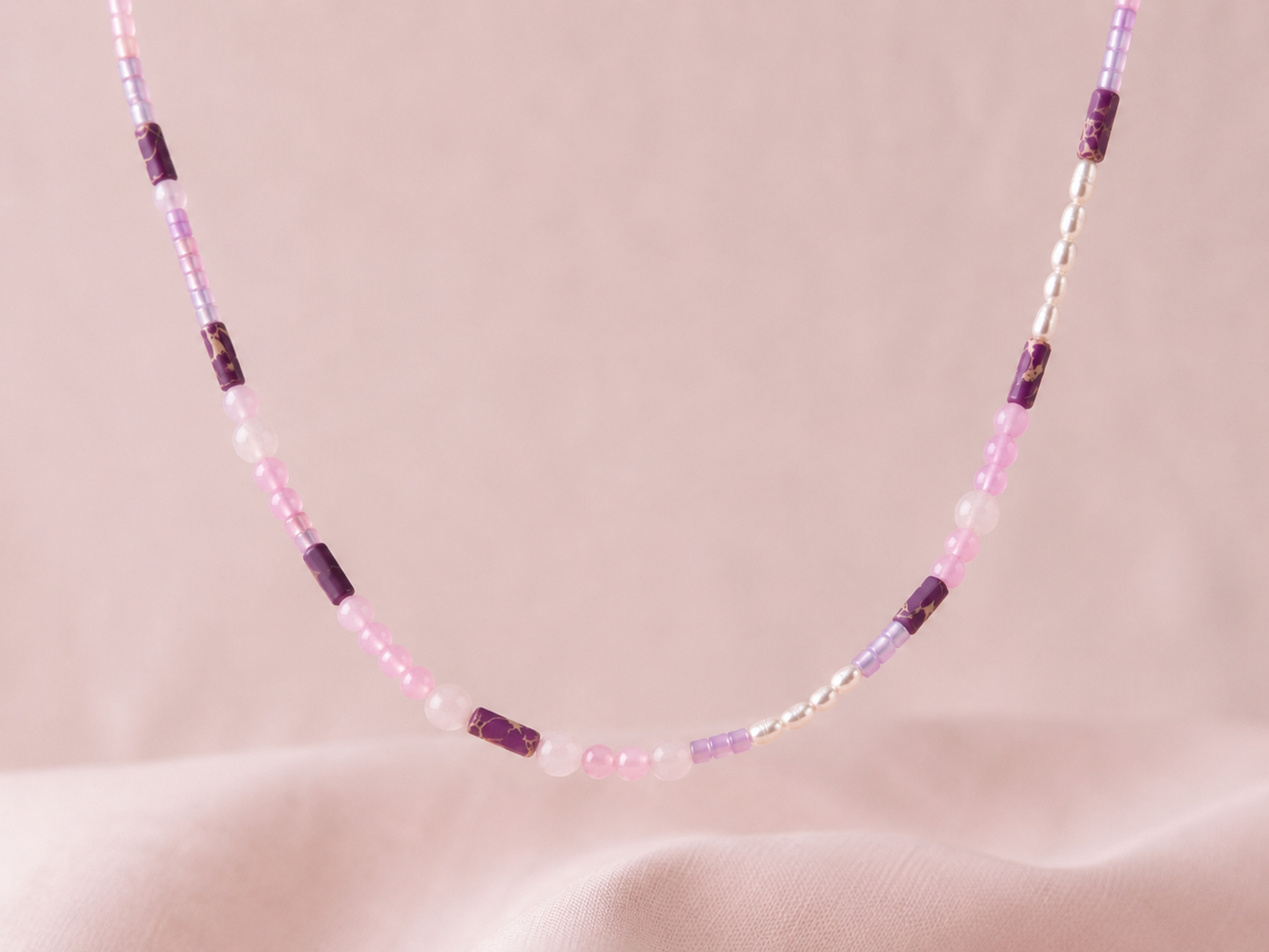 Primer plano del collar Malva sobre fondo de tela rosa, elaborado con cuentas en tonos malva y rosa, detalles morados y pequeñas piezas de acero inoxidable.