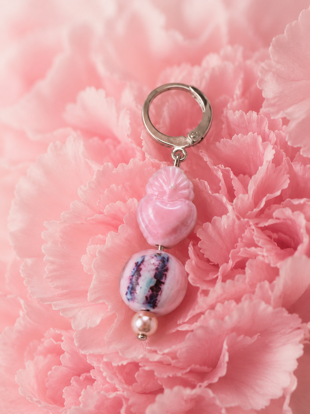 Primer plano de un pendiente artesanal Cuore con aro de acero inoxidable plateado, corazón rosa tallado, cuenta redonda veteada en tonos rosa, blanco y ciruela, y perla de agua dulce sobre un clavel rosa.