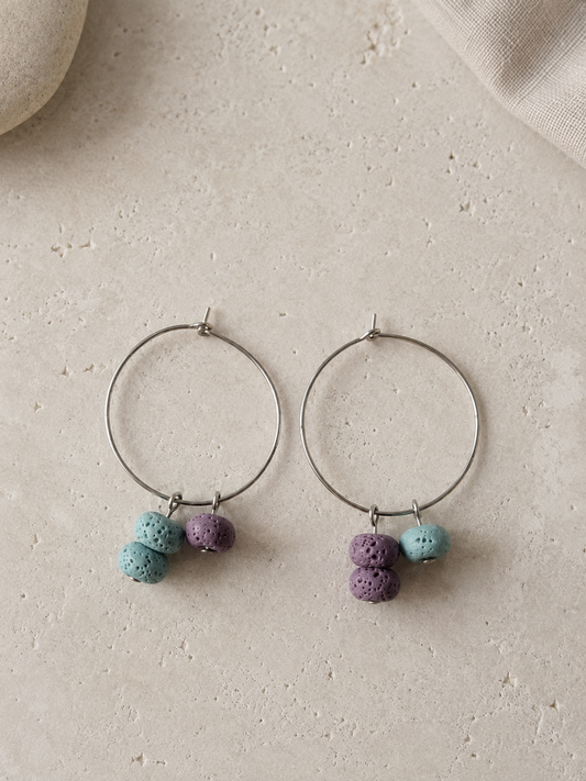 Pendientes artesanales Arrecife con aros de acero inoxidable plateado y cuentas de piedra volcánica porosa en tonos turquesa y lila, formando un diseño asimétrico sobre fondo de piedra clara.