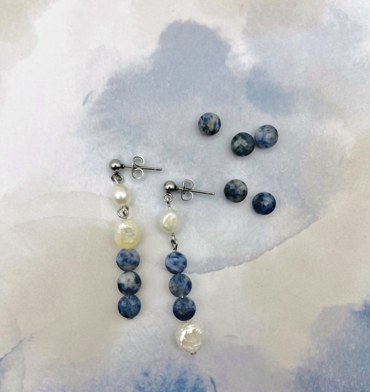 Pendientes Bajamar de Cuadratura Studio sobre fondo acuarela azul: pendiente largo asimétrico con botón plateado, perlas barrocas blancas y tres cuentas de sodalita azul mateada, junto a pendientes de tuerca de sodalita azul. Montados a mano en Murcia.`