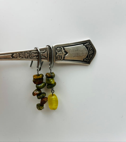 Pendientes Elena – dos aros plateados con chips de unakita verde oliva y marrón apilados y cuenta ovalada de ámbar amarillo natural colgando, colgados del mango de un cubierto de plata antigua labrada con motivos florales, fondo blanco, hechos a mano en Murcia