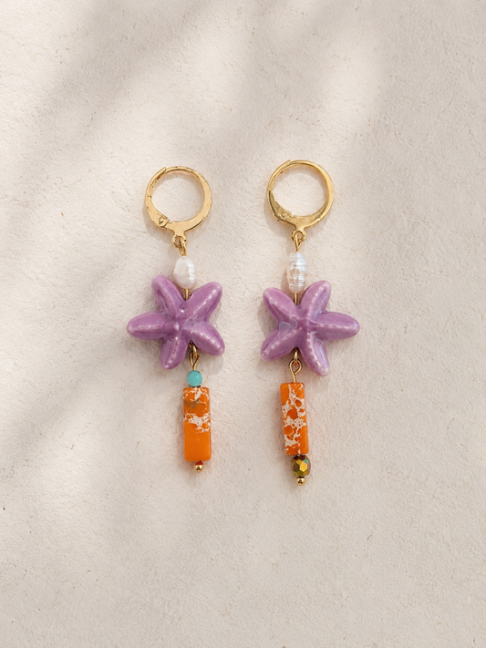 Pendientes artesanales Mediterráneo con aros de acero inoxidable chapados en oro, estrella de mar de cerámica esmaltada en color lila, perla de agua dulce y barras de jaspe sedimentario naranja, en diseño asimétrico sobre fondo de piedra clara.