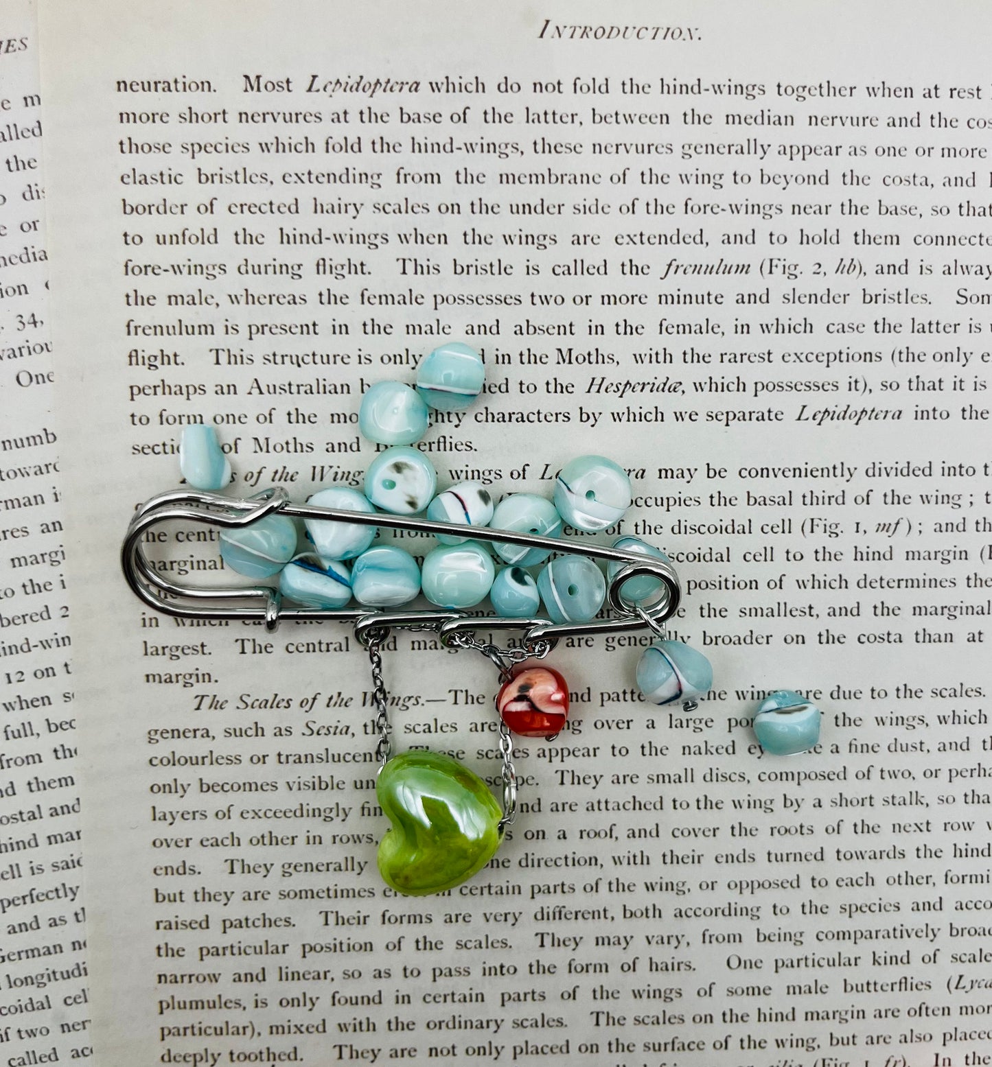 Broche Calma – imperdible plateado con semillas de rocalla azul celeste y charms de corazón de cerámica rojo y verde, hecho a mano en Murcia