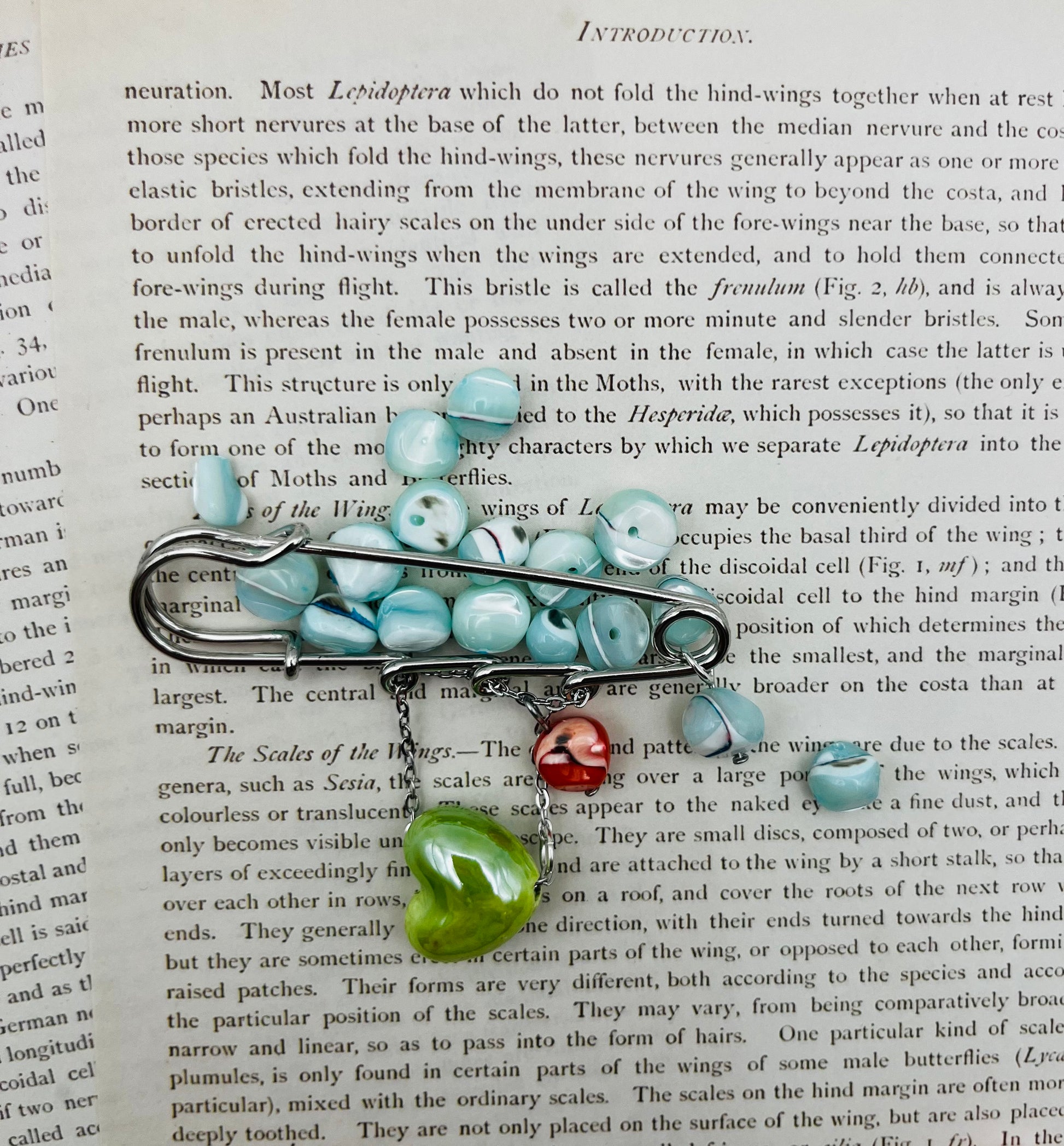 Broche Calma – imperdible plateado con semillas de rocalla azul celeste y charms de corazón de cerámica rojo y verde, hecho a mano en Murcia