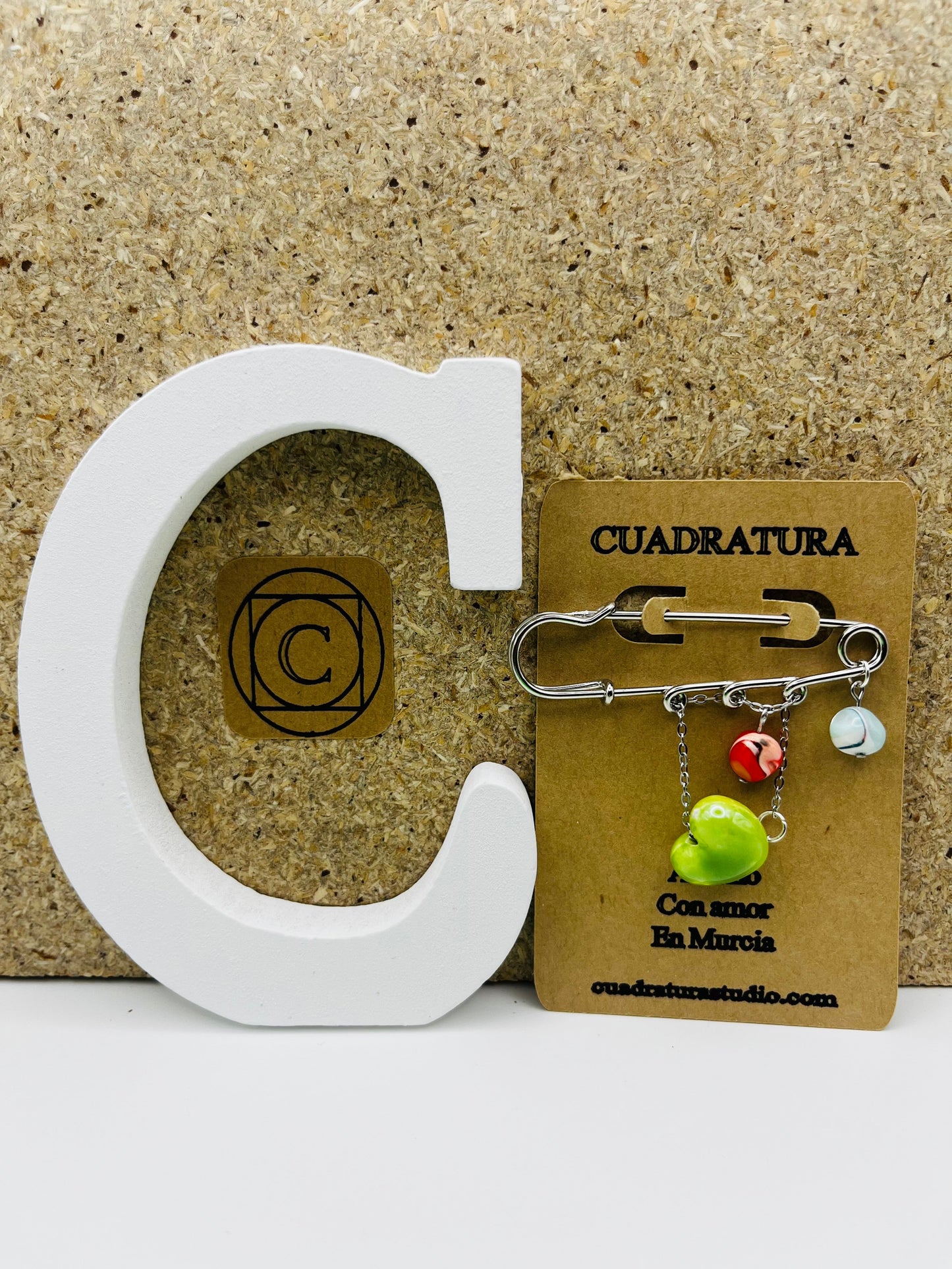 Broche Calma de Cuadratura Studio con imperdible plateado y colgantes de howlita verde, millefiori rojo y corazón de cerámica, en tarjeta kraft junto a letra C blanca, montado a mano en Murcia.