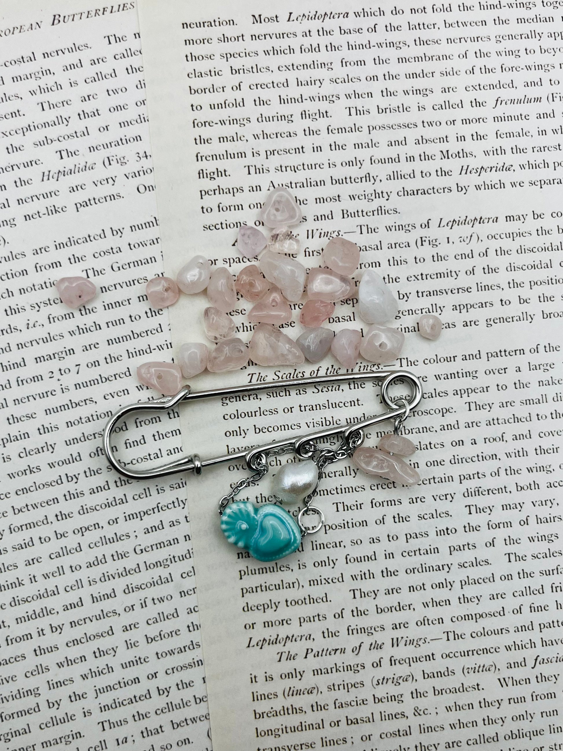 Vista cenital del Broche Cuore en imperdible plateado con corazón de cerámica turquesa, perla blanca y chip de cuarzo rosa, sobre libro vintage rodeado de chips de cuarzo rosa sueltos, montado a mano en Murcia
