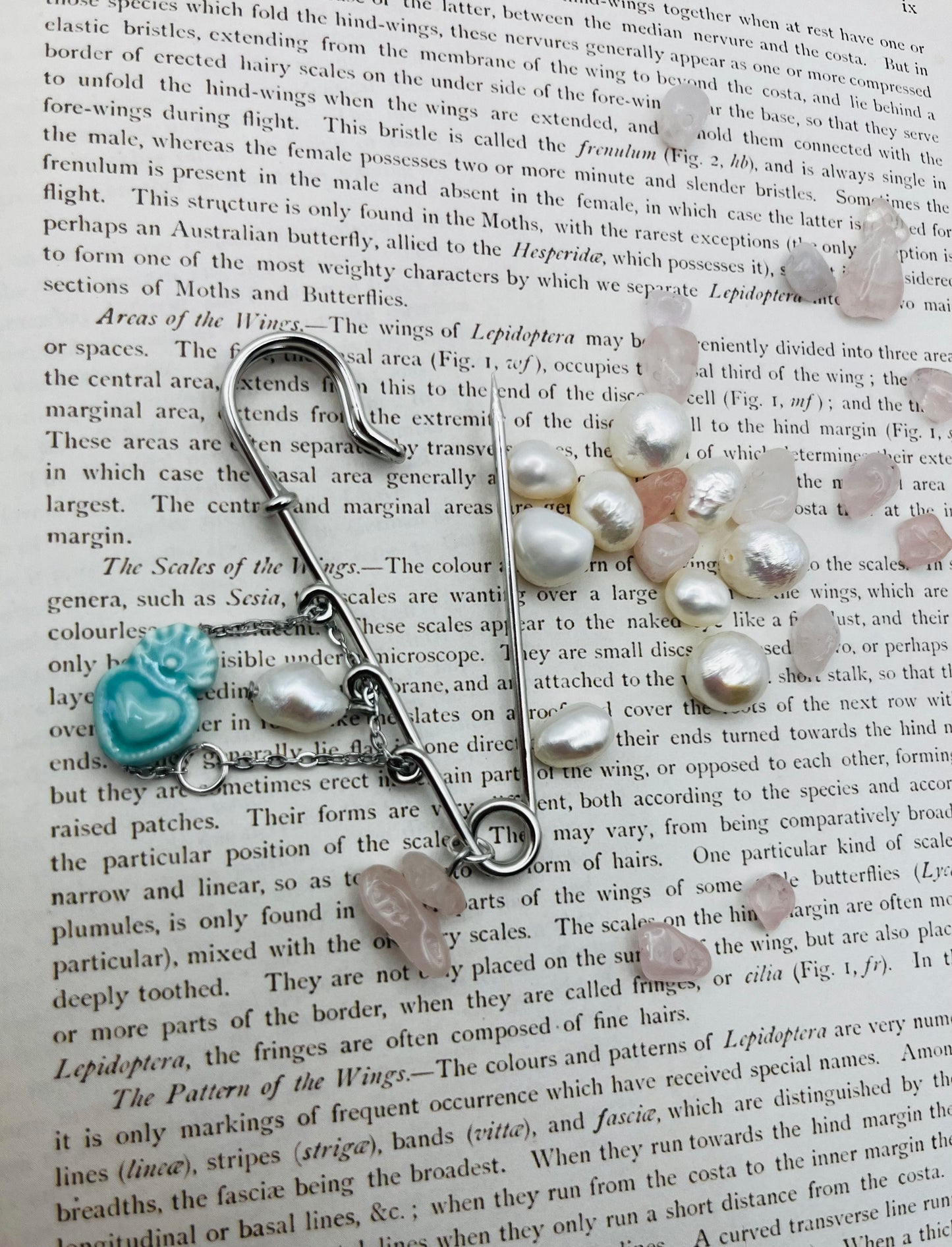 Detalle del Broche Cuore en imperdible plateado con corazón de cerámica turquesa, perla blanca y chip de cuarzo rosa, sobre libro vintage rodeado de chips de cuarzo rosa y perlas barrocas sueltas, montado a mano en Murcia
