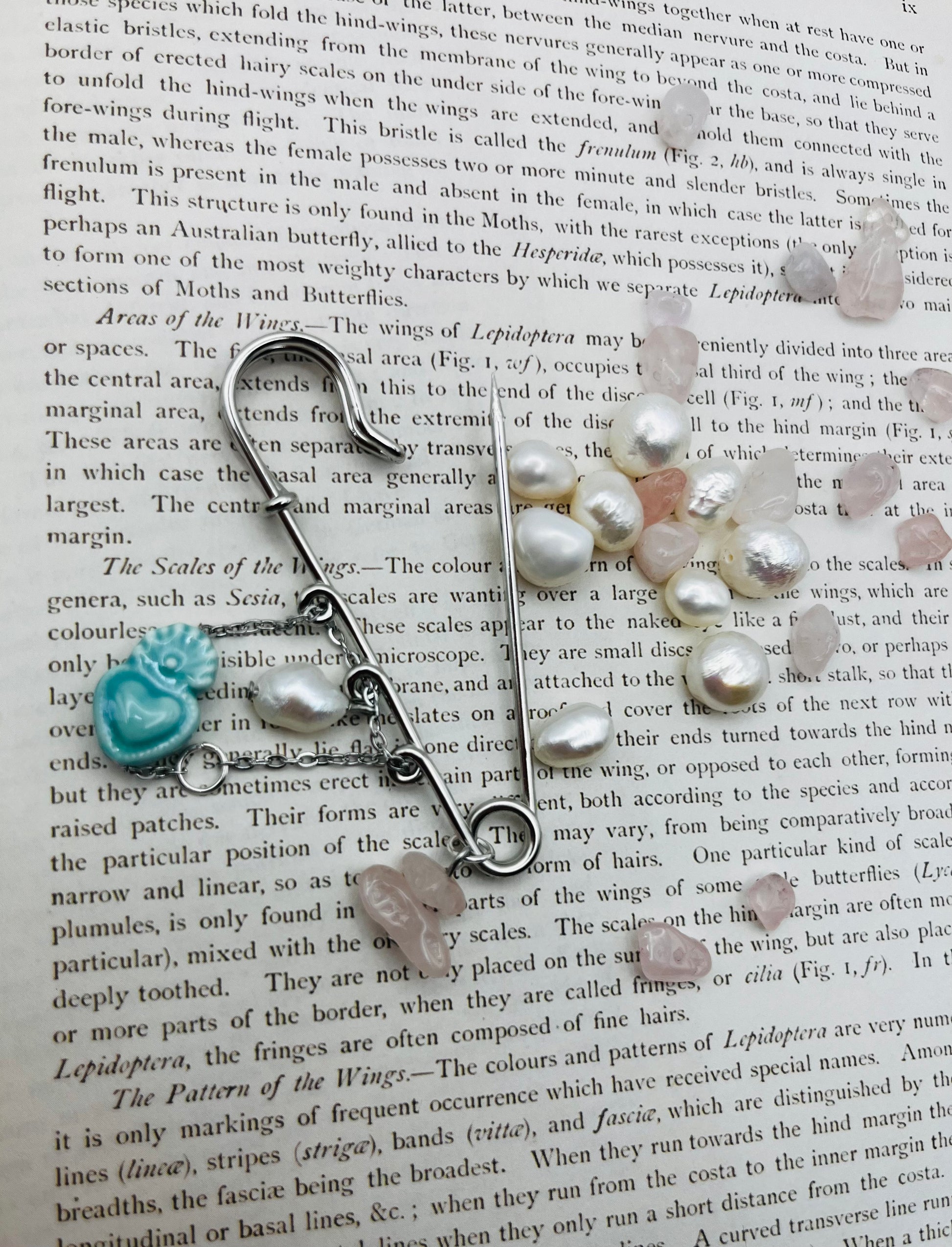 Detalle del Broche Cuore en imperdible plateado con corazón de cerámica turquesa, perla blanca y chip de cuarzo rosa, sobre libro vintage rodeado de chips de cuarzo rosa y perlas barrocas sueltas, montado a mano en Murcia
