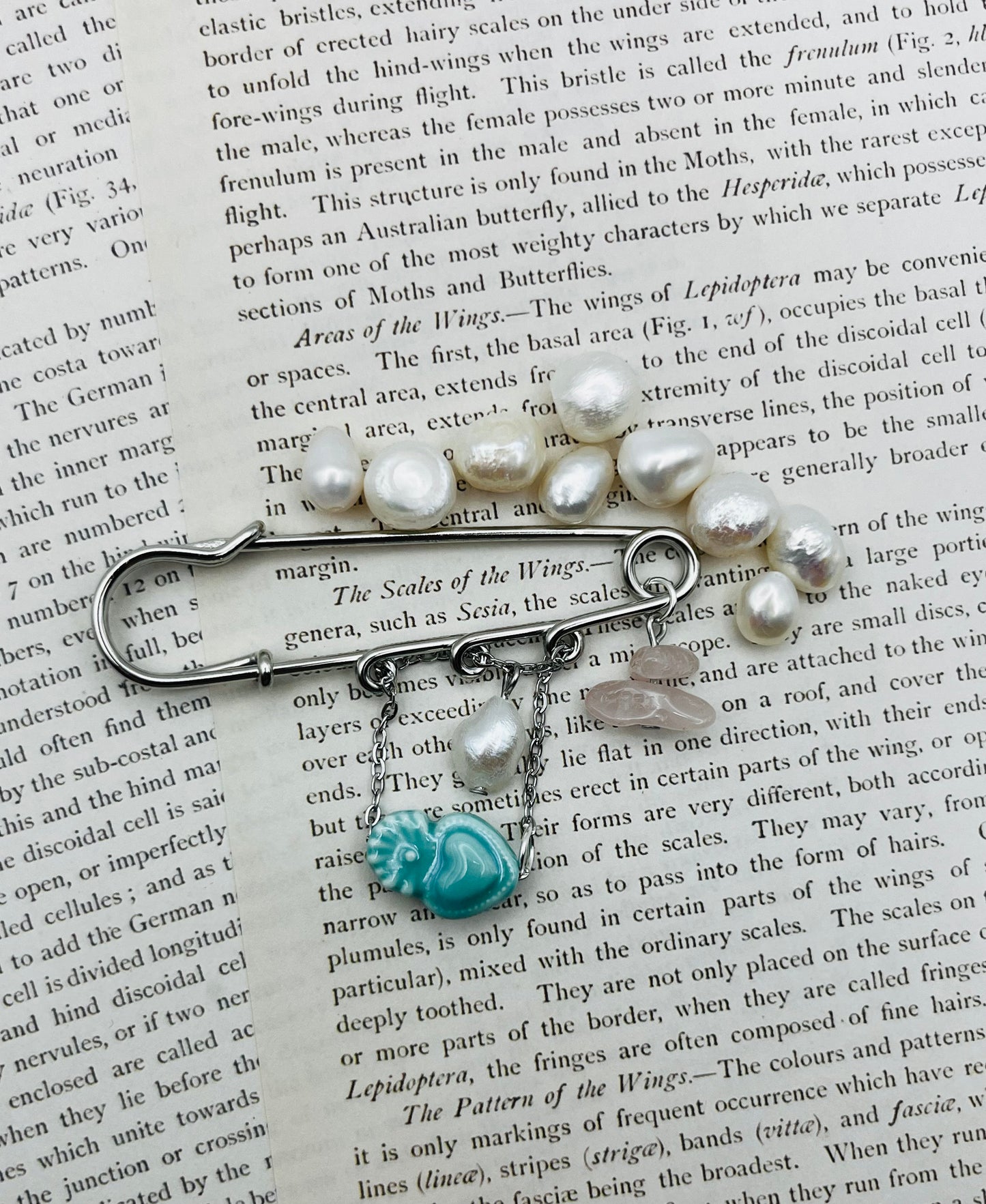 Vista cenital del Broche Cuore en imperdible plateado con corazón de cerámica turquesa, perla blanca y chip de cuarzo rosa, sobre libro vintage de entomología rodeado de perlas barrocas sueltas, montado a mano en Murcia
