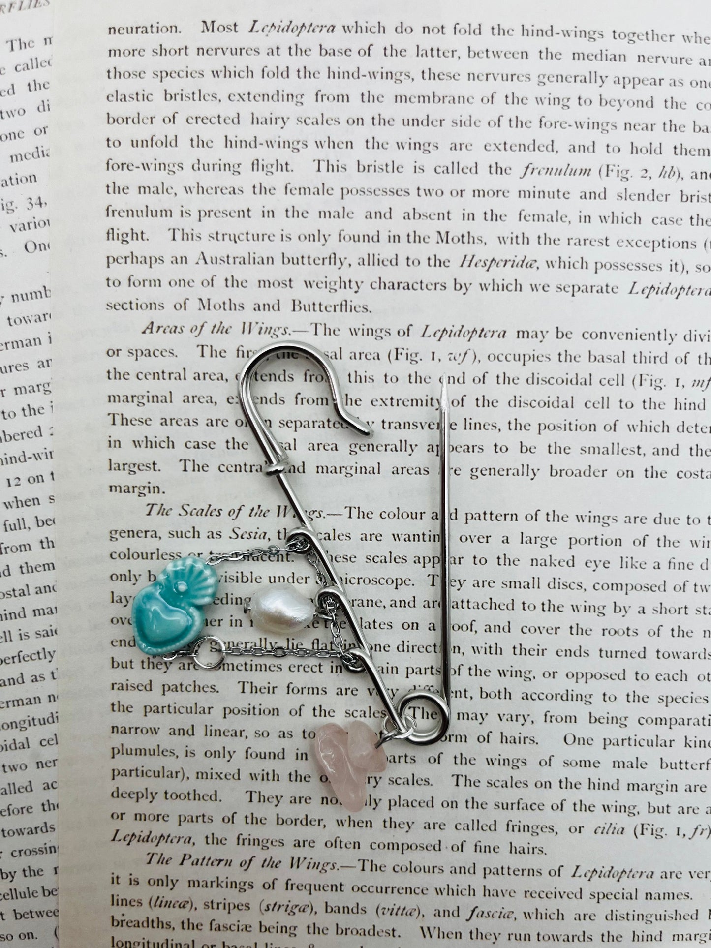 Broche Cuore en imperdible plateado con corazón de cerámica turquesa, perla irregular blanca y chip de cuarzo rosa, sobre página de libro vintage de entomología con perlas sueltas, montado a mano en Murcia
