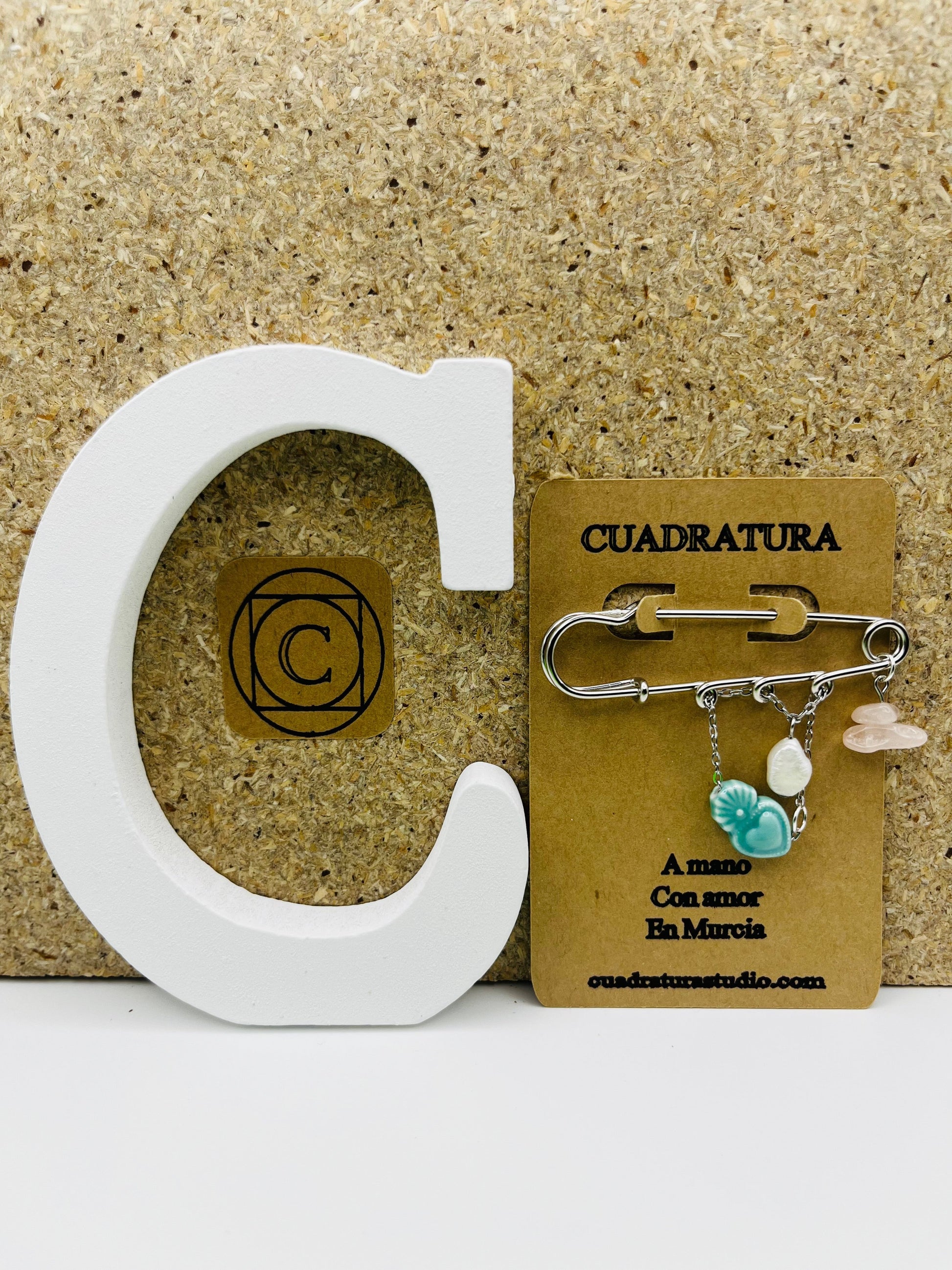 Broche Cuore en imperdible plateado con corazón de cerámica turquesa, perla irregular blanca y chip de cuarzo rosa, presentado en tarjeta Cuadratura junto a letra C blanca, montado a mano en Murcia
