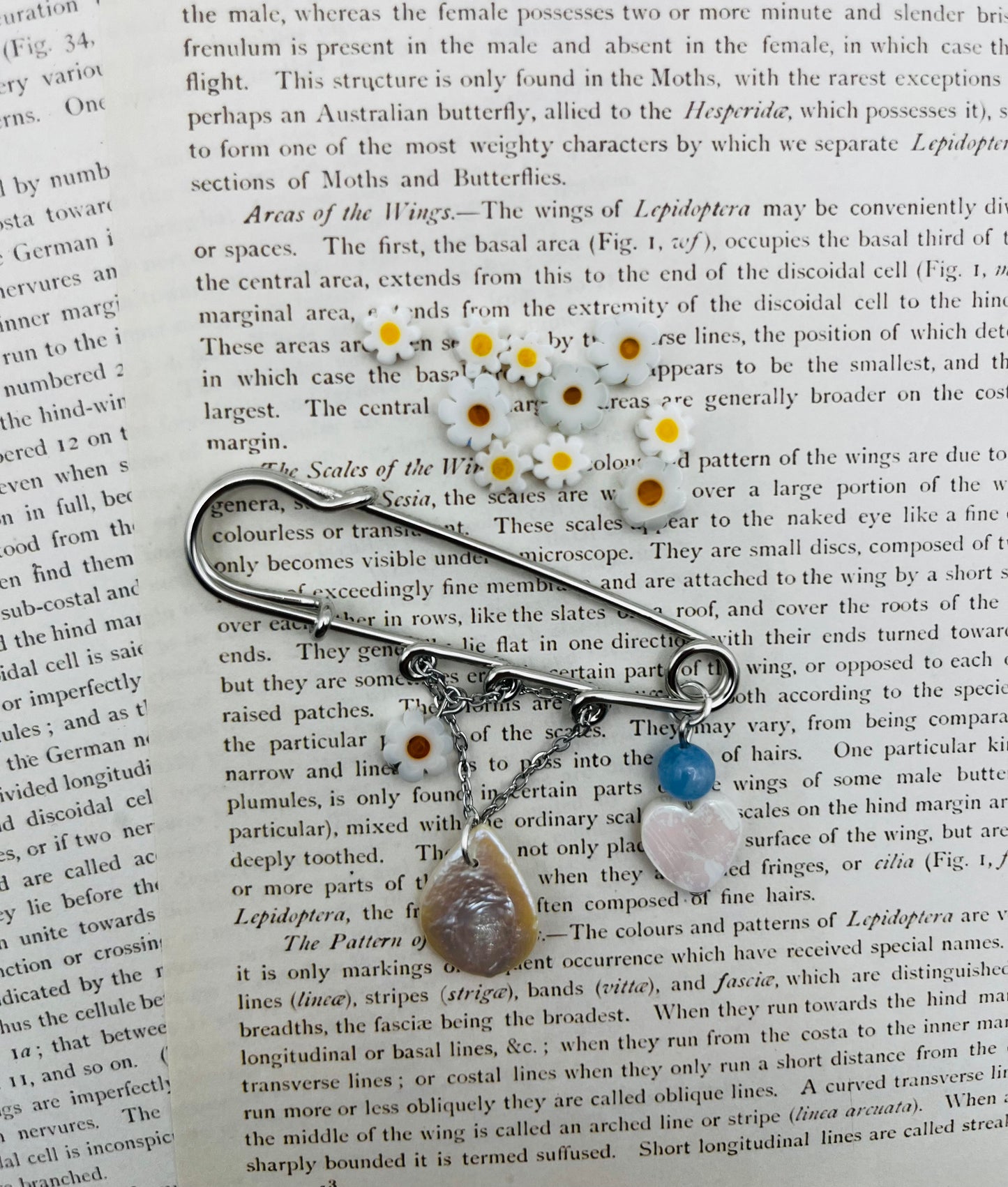 Broche Periwinkle – imperdible plateado con flor de margarita, cuenta azul, corazón de nácar y pieza gota de nácar, rodeado de flores de margarita, hecho a mano en Murcia