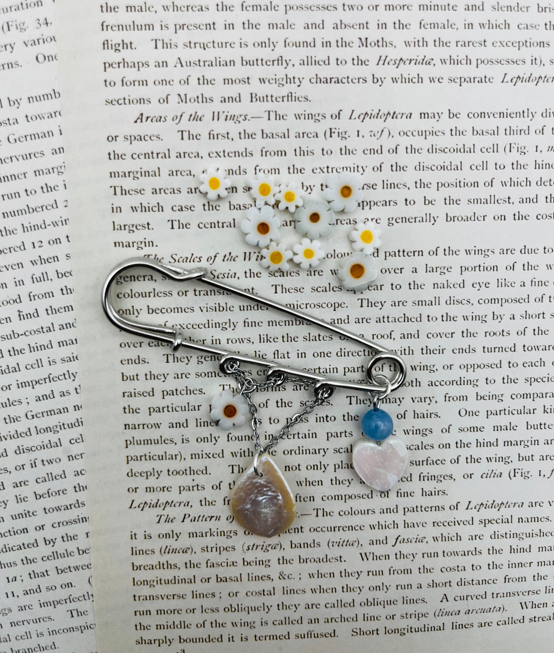 Broche Periwinkle – imperdible plateado con flor de margarita, cuenta azul, corazón de nácar y pieza gota de nácar, rodeado de flores de margarita, hecho a mano en Murcia