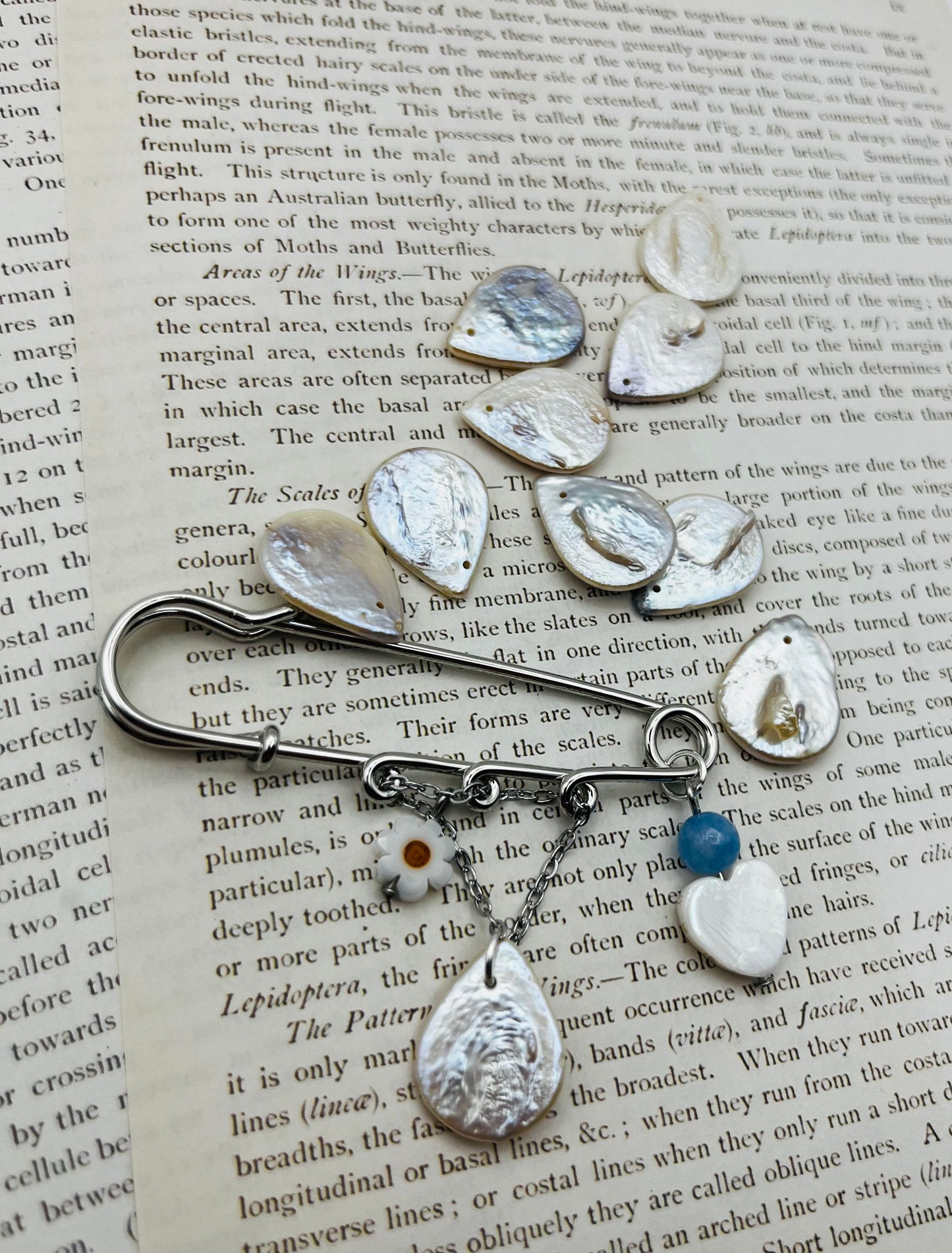 Broche Periwinkle – imperdible plateado con flor de margarita, cuenta azul, corazón de nácar y pieza gota de nácar, rodeado de piezas de nácar sueltas, hecho a mano en Murcia