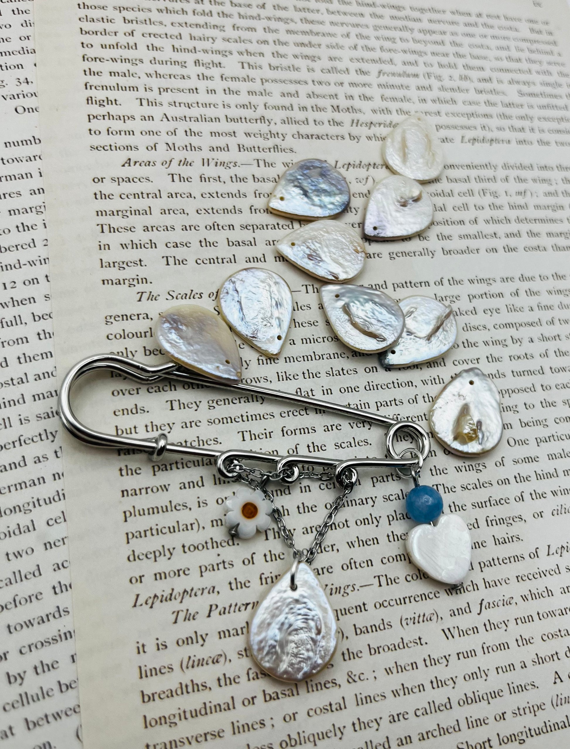 Broche Periwinkle – imperdible plateado con flor de margarita, cuenta azul, corazón de nácar y pieza gota de nácar, rodeado de piezas de nácar sueltas, hecho a mano en Murcia