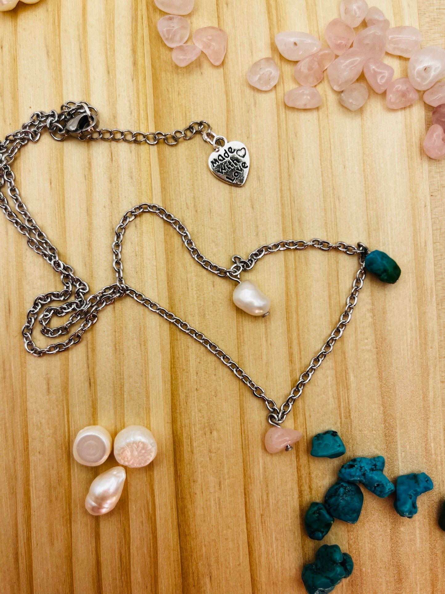 Detalle del colgante asimétrico Brisa con chip de turquesa azul, perla barroca blanca y chip de cuarzo rosado en cadena plateada con charm "Made with Love", rodeado de materiales sueltos, montado a mano en Murcia
