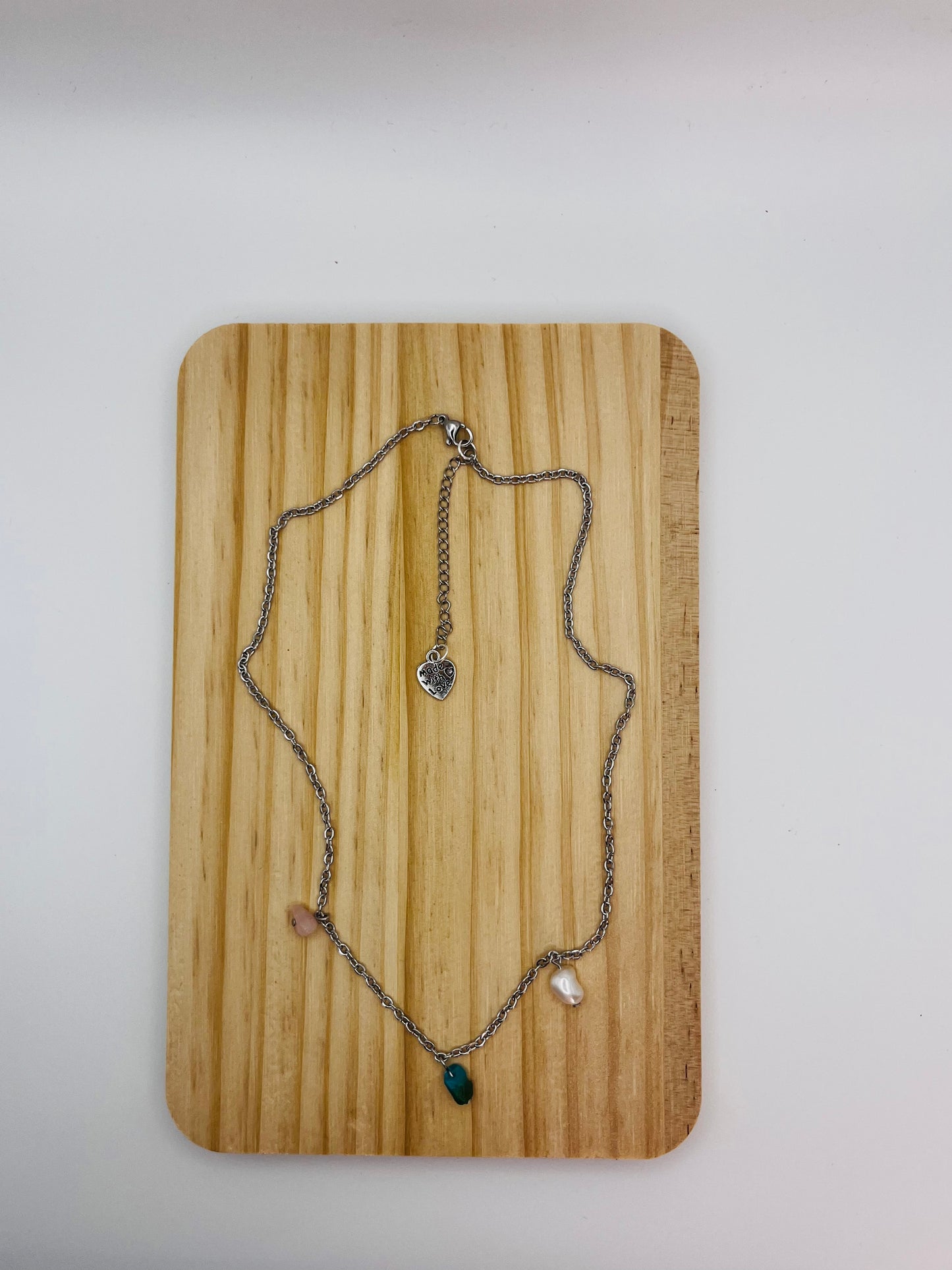 Vista completa del colgante asimétrico Brisa con chip de turquesa natural, perla barroca blanca y chip de cuarzo transparente en cadena plateada con charm corazón, sobre tabla de madera, montado a mano en Murcia
