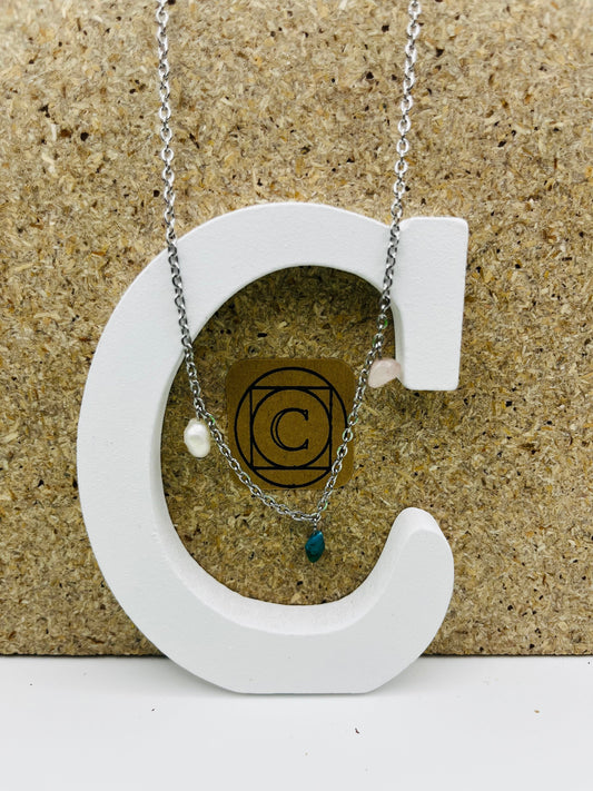 Colgante asimétrico Brisa con chip de turquesa natural, perla barroca blanca y chip de cuarzo transparente en cadena fina plateada, presentado sobre letra C blanca con etiqueta Cuadratura, montado a mano en Murcia
