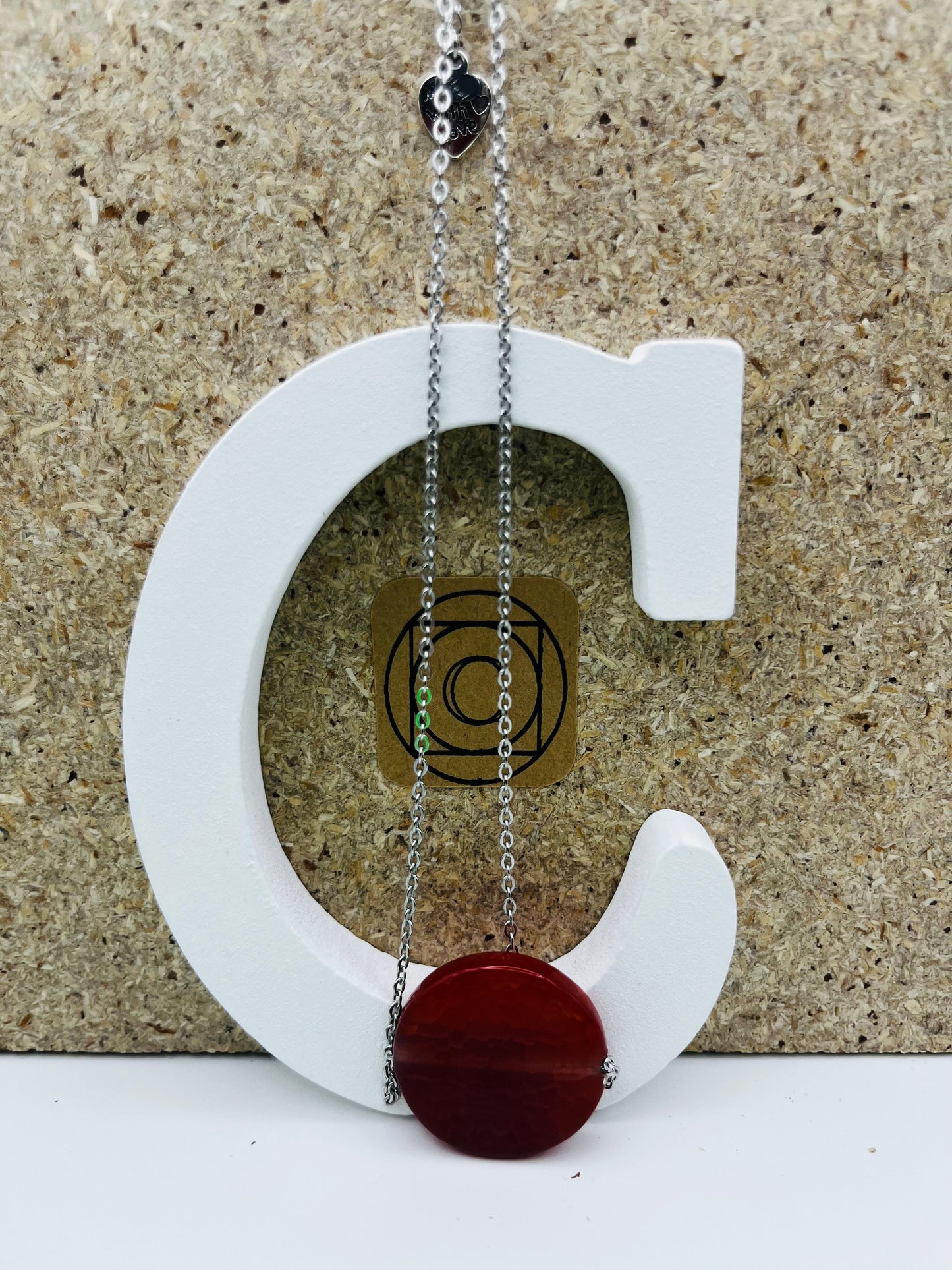 Colgante Cocedor con gran disco circular de tagua natural en rojo burdeos y charm "Made with Love" en cadena larga fina plateada, presentado sobre letra C blanca con etiqueta Cuadratura, montado a mano en Murcia
