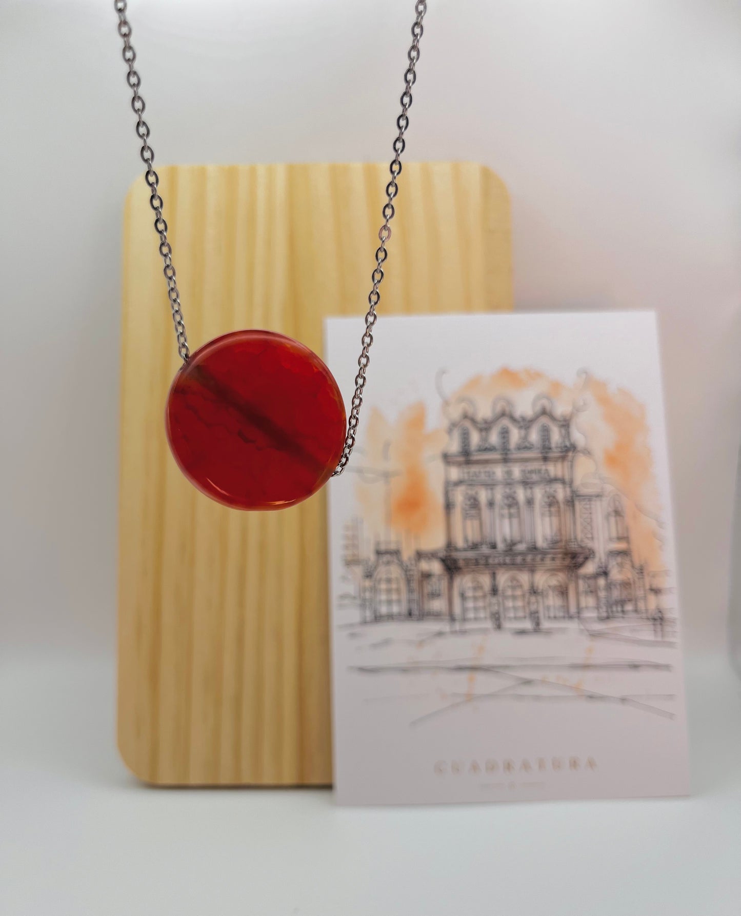 Colgante Cocedor con disco de tagua natural en rojo burdeos en cadena plateada, apoyado en soporte de madera acanalado junto a ilustración arquitectónica Cuadratura, montado a mano en Murcia
