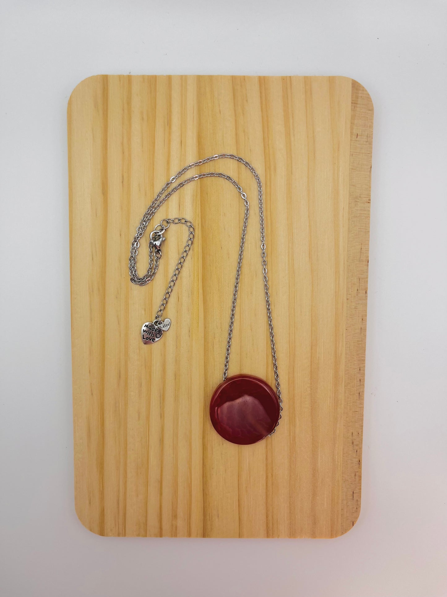 Vista completa del colgante Cocedor con disco circular de tagua natural en rojo burdeos y charm "Made with Love" en cadena larga fina plateada, sobre tabla de madera, montado a mano en Murcia
