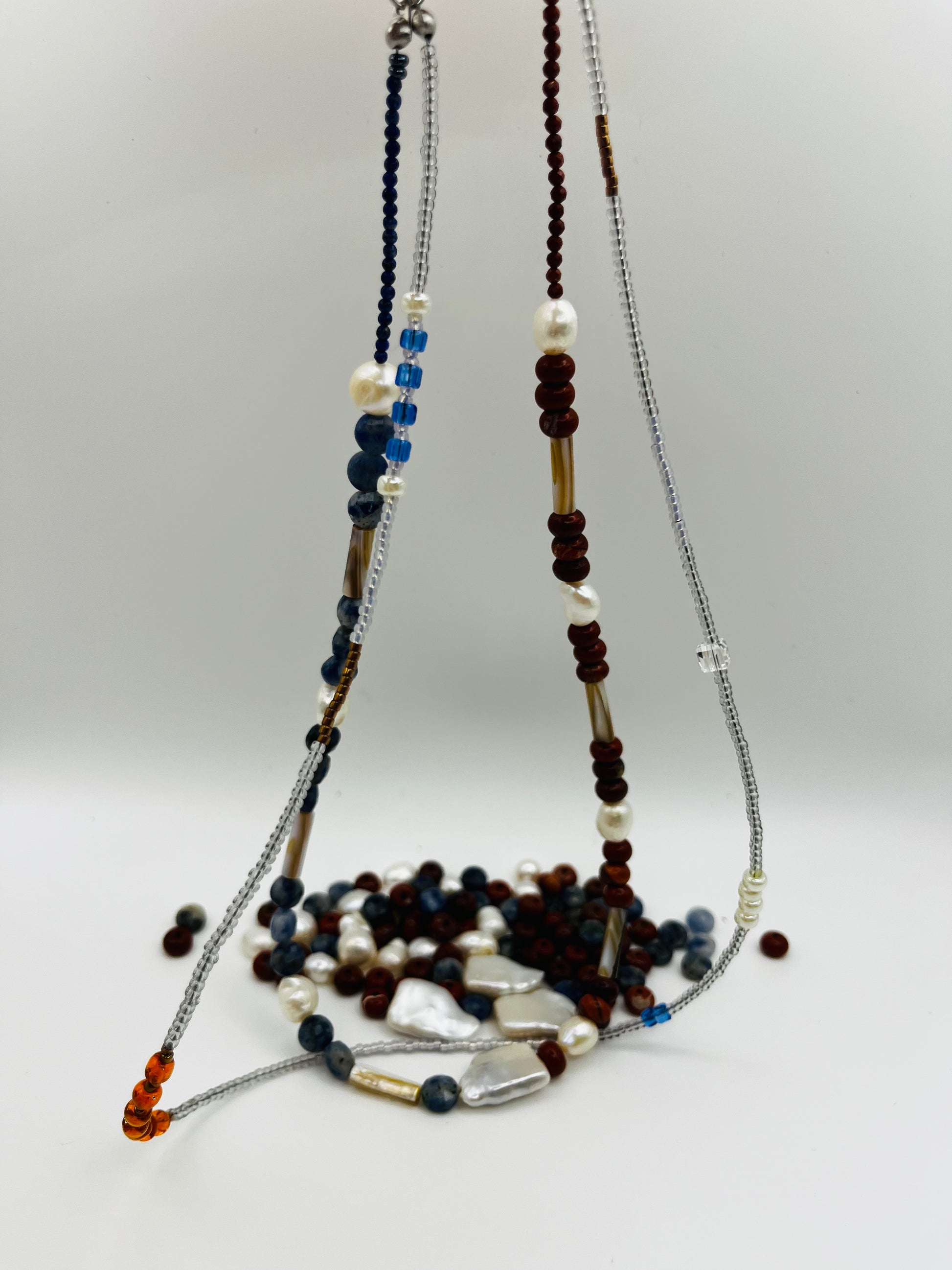 Collar Bajamar – doble collar colgado con jaspe rojo, lapislázuli azul facetado, perlas barrocas blancas, cuentas tubulares plateadas y semillas de rocalla ámbar naranja, con cuentas sueltas de jaspe y lapislázuli en el fondo, hecho a mano en Murcia