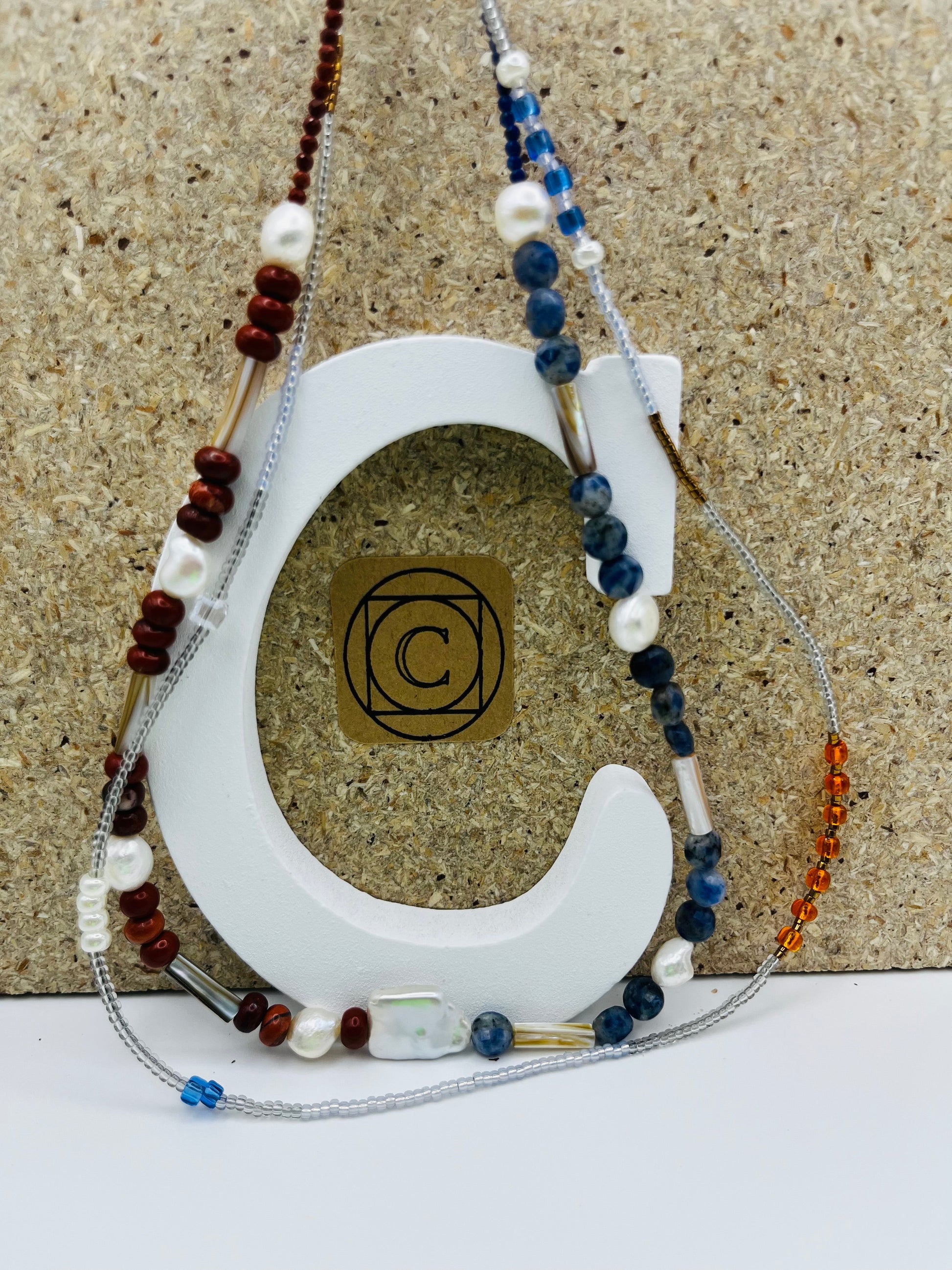 Collar Bajamar – doble collar con jaspe rojo, lapislázuli azul, perlas barrocas blancas, cuentas tubulares plateadas y semillas de rocalla transparentes, azules y ámbar, sobre letra C blanca con logo Cuadratura, hecho a mano en Murcia