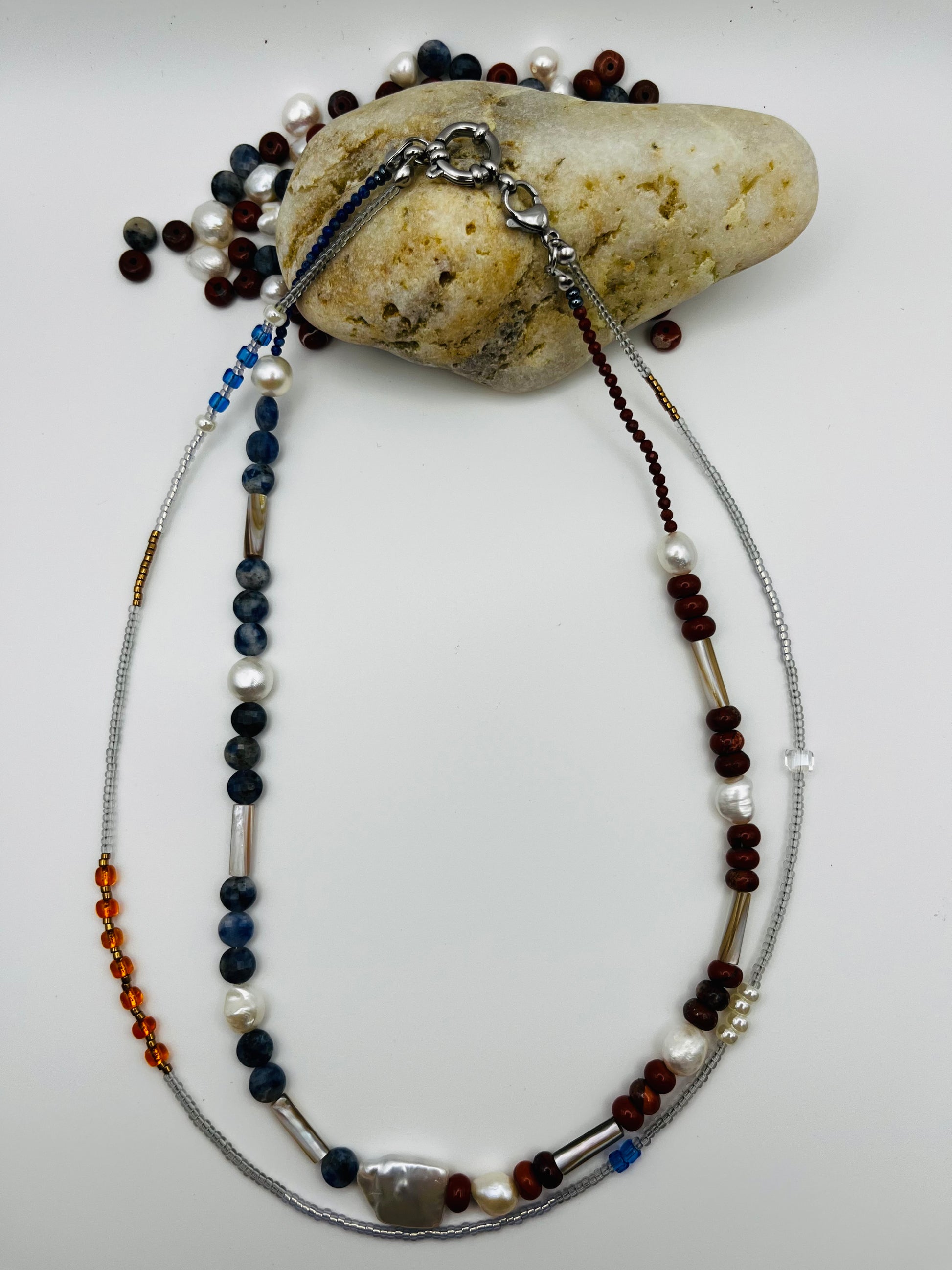 Collar Bajamar – doble collar con jaspe rojo, lapislázuli azul facetado, perlas barrocas blancas, cuentas tubulares plateadas y semillas de rocalla, apoyado sobre piedra de playa con cuentas sueltas de jaspe, lapislázuli y perlas, hecho a mano en Murcia
