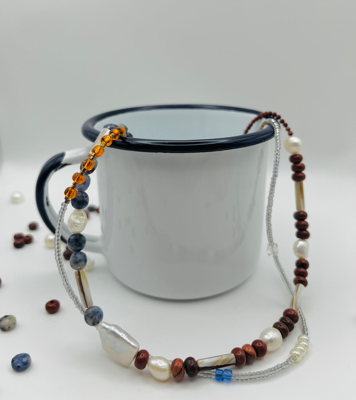 Collar Bajamar – doble collar con jaspe rojo, lapislázuli azul facetado, perlas barrocas blancas, cuentas tubulares plateadas y semillas de rocalla ámbar naranja, sobre taza esmaltada blanca con borde negro, hecho a mano en Murcia
