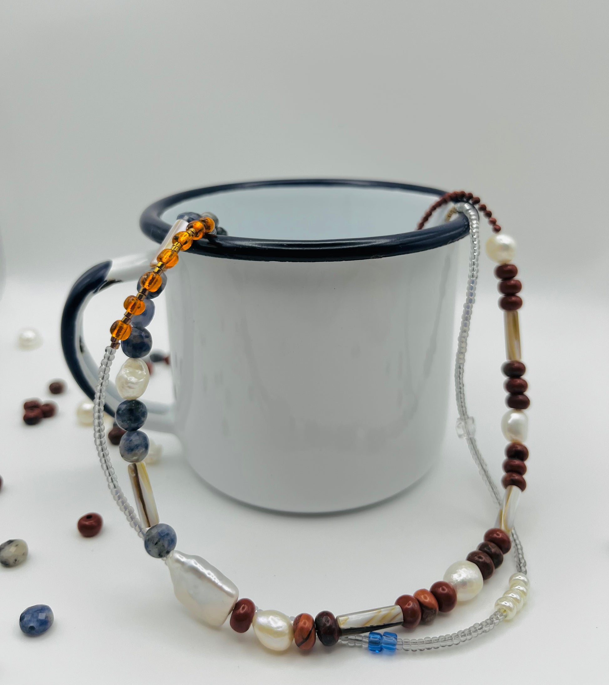 Collar Bajamar – doble collar con jaspe rojo, lapislázuli azul facetado, perlas barrocas blancas, cuentas tubulares plateadas y semillas de rocalla ámbar naranja, sobre taza esmaltada blanca con borde negro, hecho a mano en Murcia