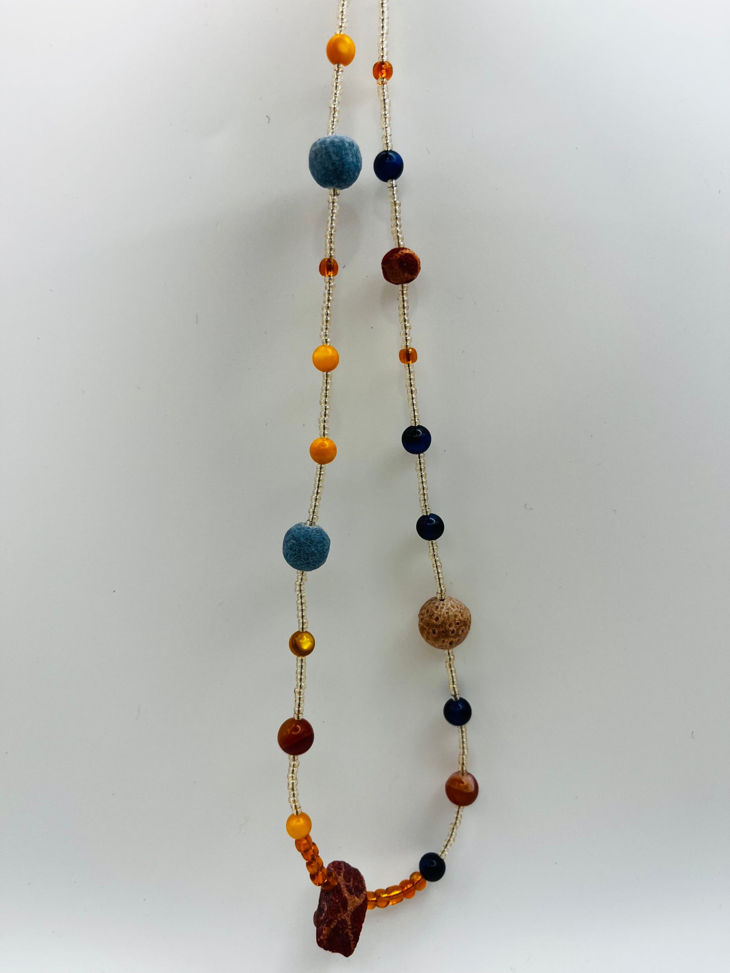 Collar Cala – collar largo con semillas de rocalla transparentes, lava volcánica azul y terracota, ámbar naranja, lapislázuli azul marino y coral rojo natural, hecho a mano en Murcia
