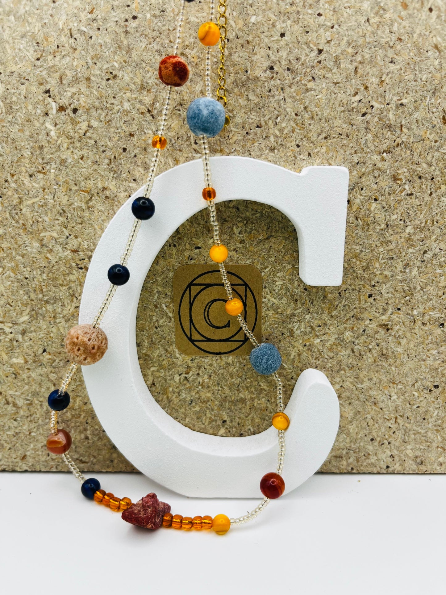 Collar Cala – collar largo con semillas de rocalla transparentes, lava volcánica azul y terracota, ámbar naranja, lapislázuli azul marino y coral rojo natural, sobre letra C blanca con logo Cuadratura, hecho a mano en Murcia