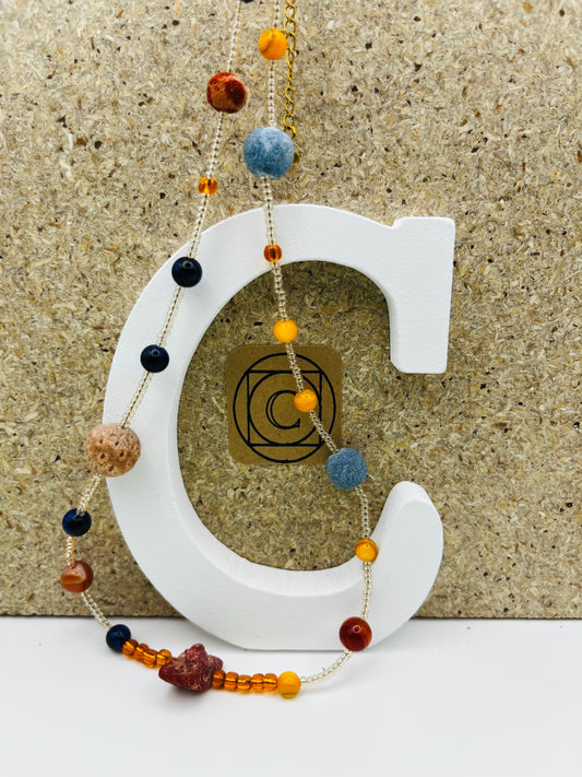 Collar Cala – collar largo con semillas de rocalla transparentes, lava volcánica azul y terracota, ámbar naranja, lapislázuli azul marino y coral rojo natural, sobre letra C blanca con logo Cuadratura, hecho a mano en Murcia