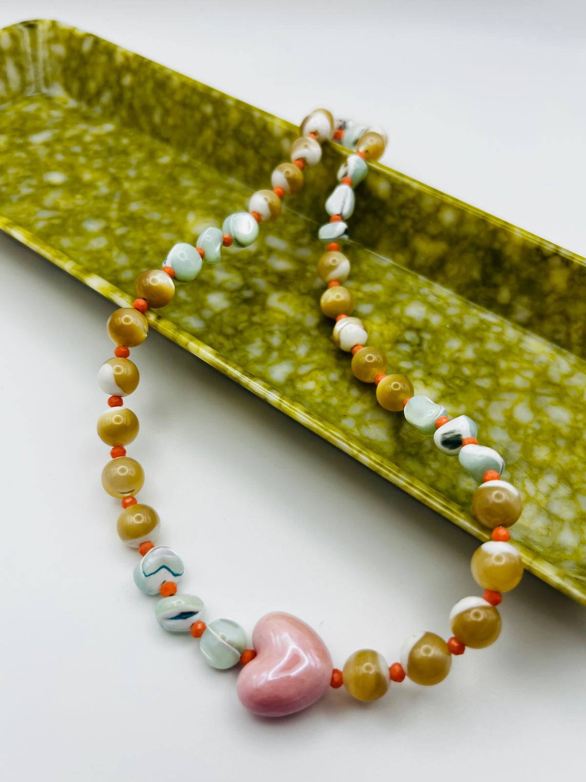 Collar Calma en ángulo sobre bandeja rectangular verde marmolada, con corazón central de cerámica rosa esmaltada en primer plano, chips de howlita marmolada, chips de amazonita turquesa, cuentas de coral rosa y perlas de agua dulce nacaradas, hecho a mano en Murcia