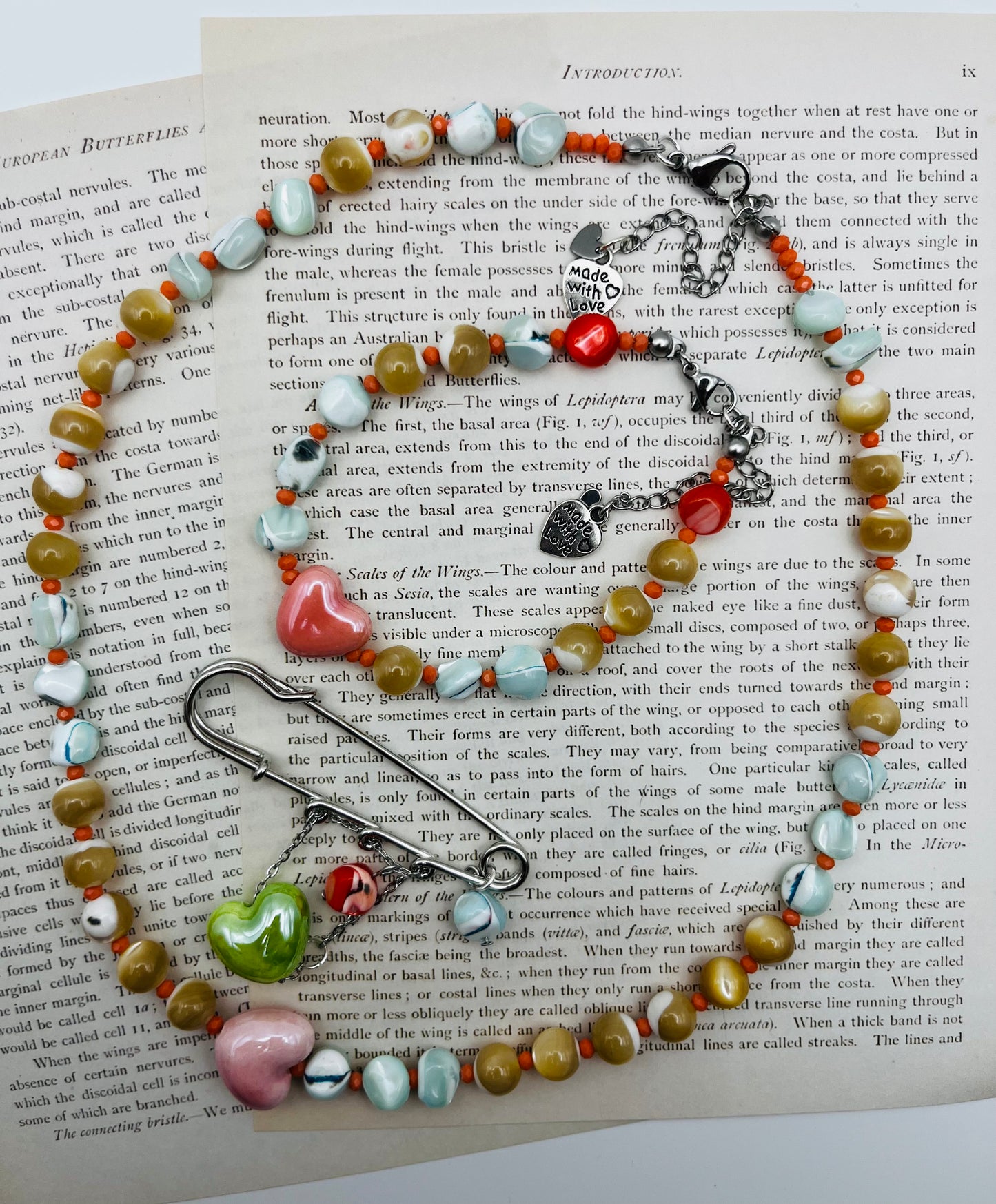 Collar y broche Calma de Cuadratura Studio sobre página de libro antiguo: collar con cuentas de cerámica, howlita, coral y corazones rosas, con dije "made with love"; broche imperdible plateado con colgantes de cerámica verde y rosa.