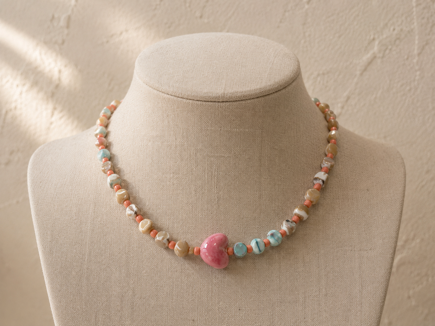 Collar Calma hecho a mano con corazón de cerámica rosa, mostrado sobre busto de joyería beige.