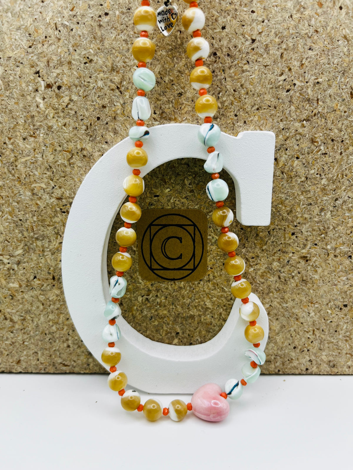 Collar artesanal Calma con cuentas de cerámica amarillo mostaza, cuentas marmoladas, semillas naranjas y colgante corazón rosa, montado a mano en Murcia
