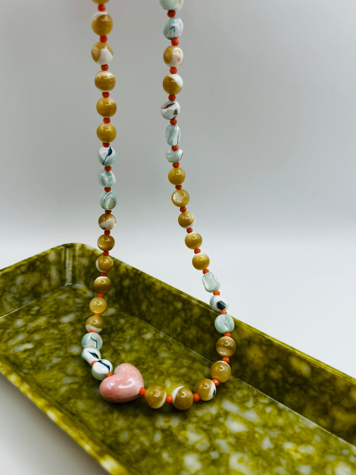 Collar Calma colgado en vertical sobre bandeja rectangular verde marmolada, mostrando los dos hilos con chips de howlita marmolada, chips de amazonita turquesa, cuentas de coral rosa y perlas de agua dulce nacaradas, con corazón central de cerámica rosa esmaltada en la base, hecho a mano en Murcia