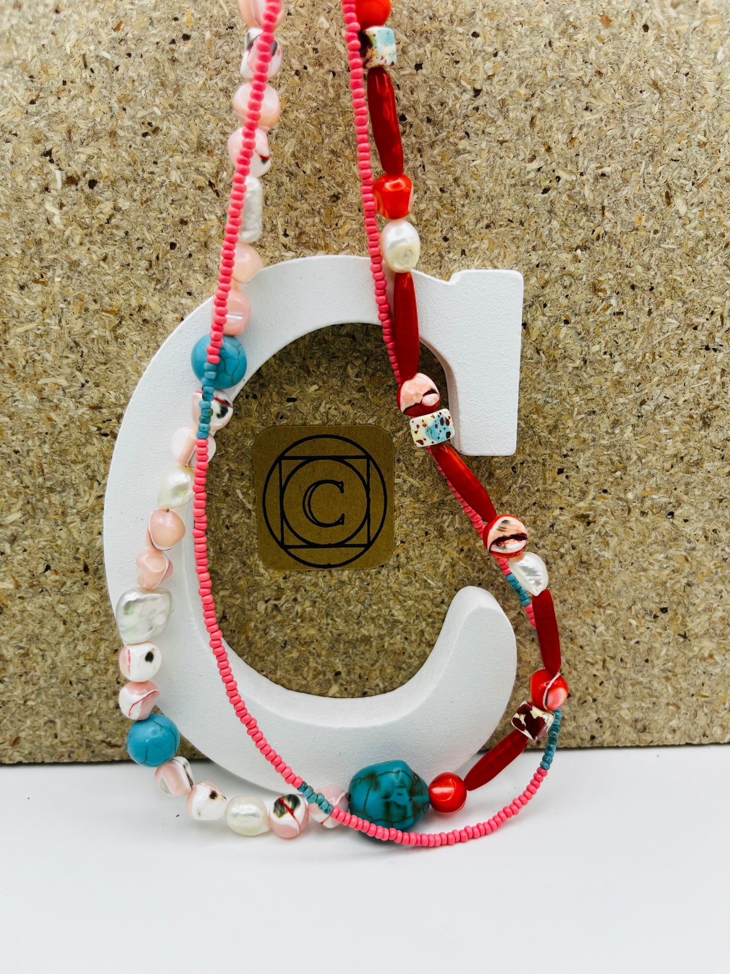 Collar Coral – doble collar con semillas de rocalla rosas, turquesa natural, coral rojo, nácar moteado, perlas barrocas blancas y cordón rojo, sobre letra C blanca con logo Cuadratura y fondo de corcho, hecho a mano en Murcia