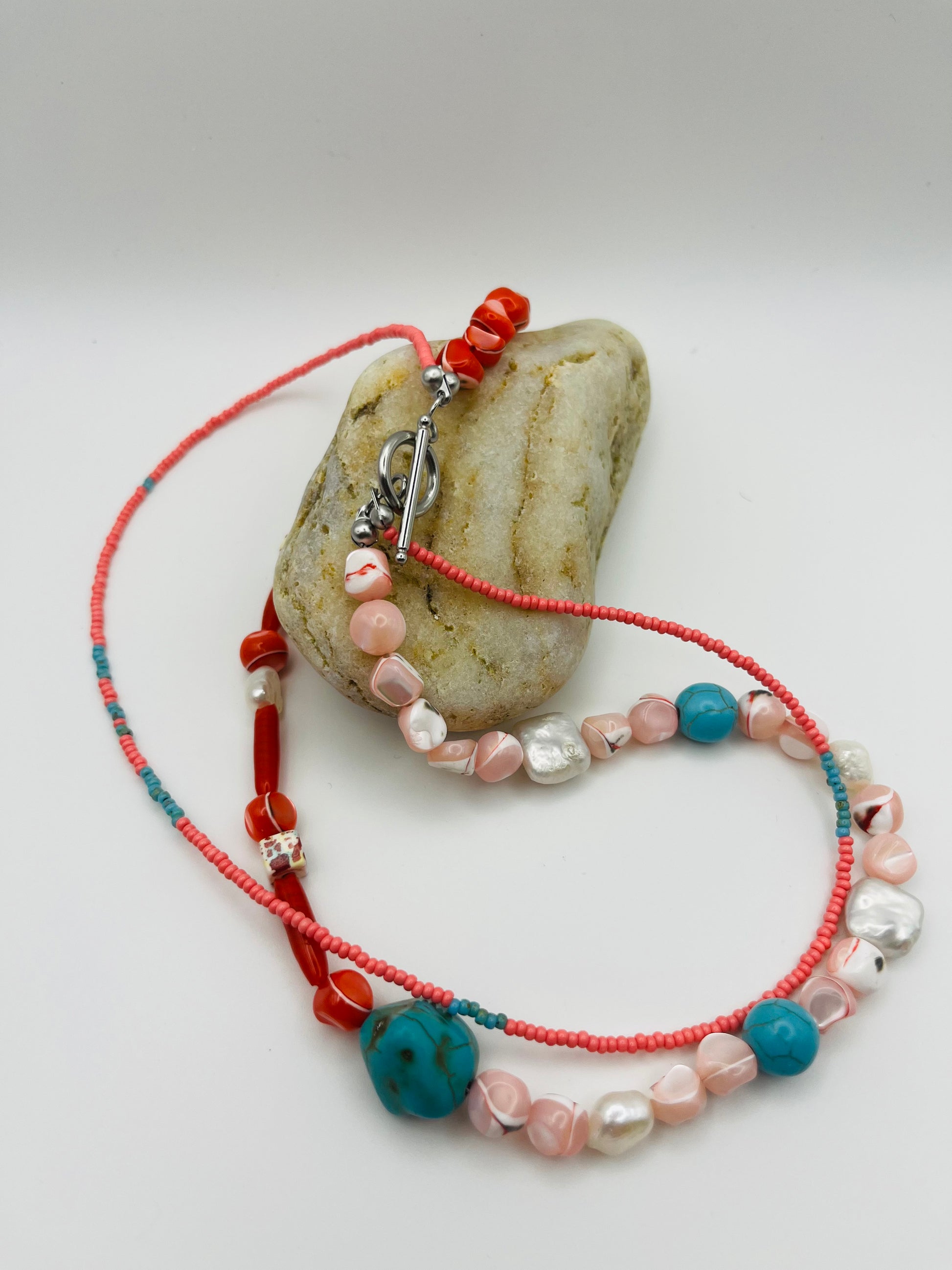 Collar Coral – detalle del doble hilo con semillas de rocalla rosas, turquesa natural, coral rojo, nácar moteado, perlas barrocas blancas y cierre toggle plateado, apoyado sobre piedra de playa, hecho a mano en Murcia