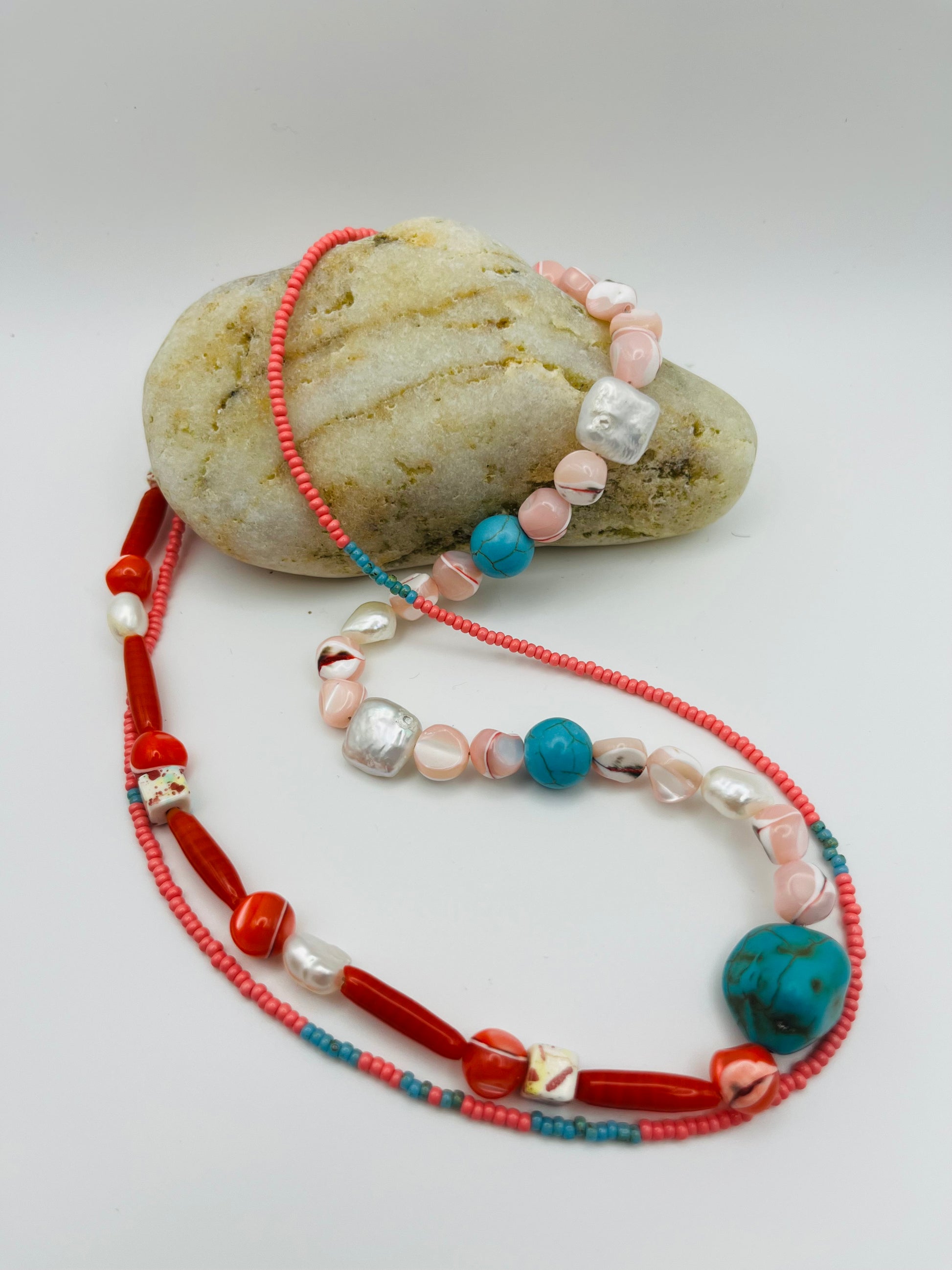 Collar Coral – vista completa del doble collar con semillas de rocalla rosas y azules, coral rojo tubular, turquesa natural, nácar moteado y perlas barrocas blancas, apoyado sobre piedra de playa, hecho a mano en Murcia