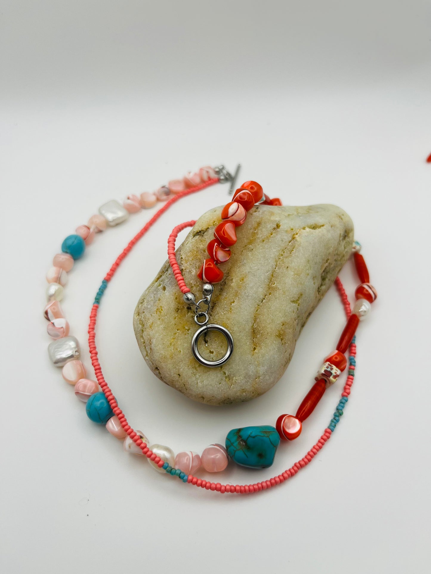 Collar Coral – doble collar con semillas de rocalla rosas, turquesa natural, coral rojo, nácar moteado y perlas barrocas blancas, con cierre toggle plateado, apoyado sobre piedra de playa, hecho a mano en Murcia