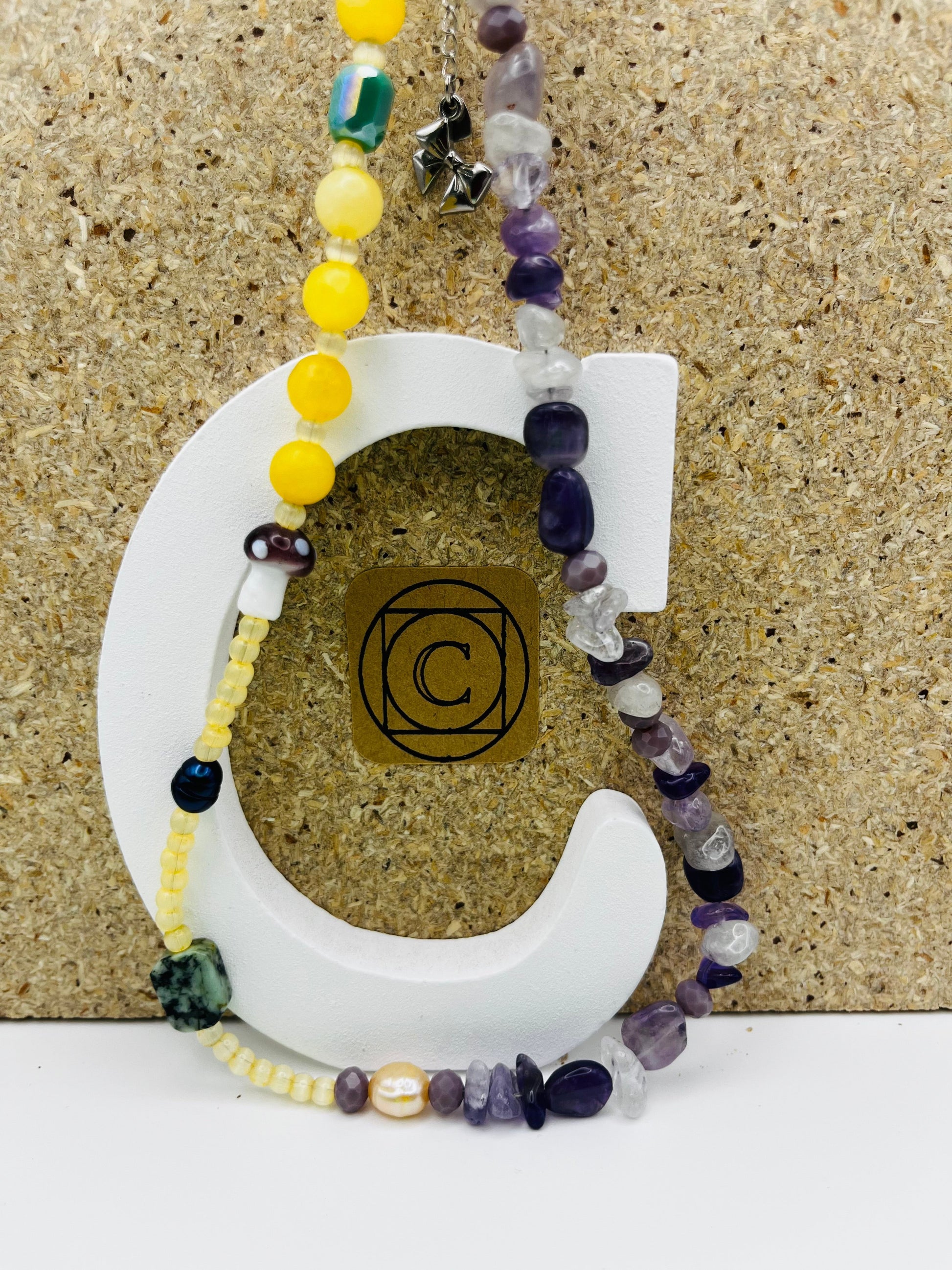 Collar Dalia – doble collar con semillas de rocalla amarillo pálido, jade amarillo, esmeralda verde, perla barroca amarilla, perla negra, seta de cristal morada, chips de amatista morada y lila, cuarzo transparente y ahumado, y cadena con charm de lazo plateado, sobre letra C blanca con logo Cuadratura, hecho a mano en Murcia

