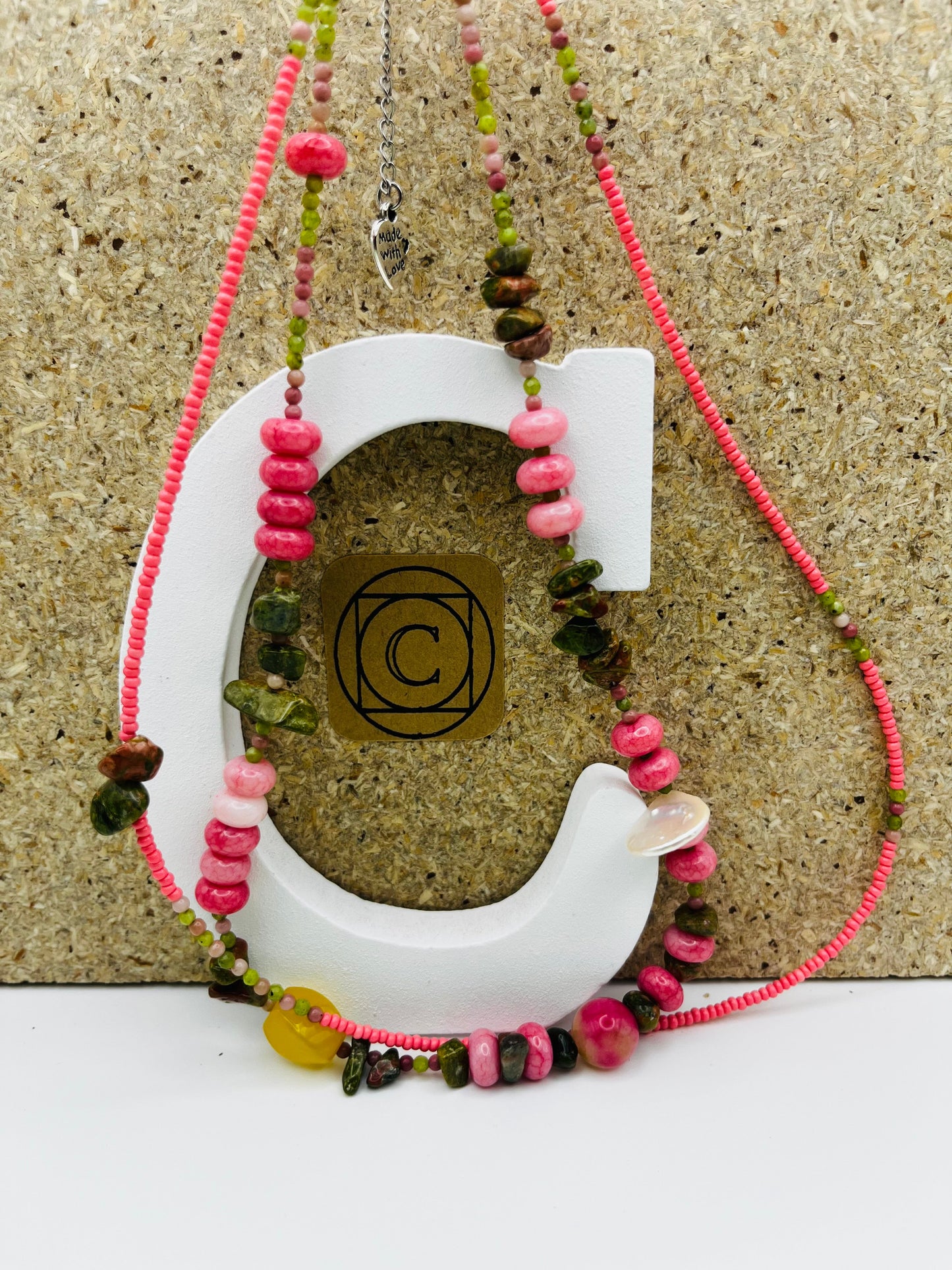 Collar Elena – doble collar con semillas de rocalla rosas, rodocrosita rosa, chips de turmalina verde y marrón, cuenta amarilla translúcida, concha de nácar blanca y charm "Made with Love" plateado, sobre letra C blanca con logo Cuadratura y fondo de corcho, hecho a mano en Murcia