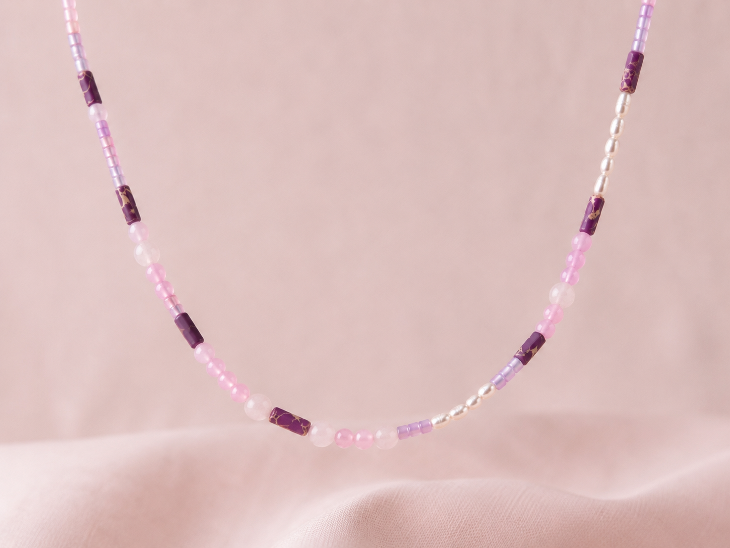 Primer plano del collar Malva sobre fondo de tela rosa, elaborado con cuentas en tonos malva y rosa, detalles morados y pequeñas piezas de acero inoxidable.