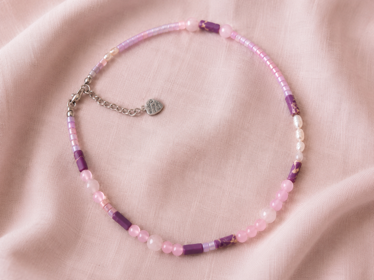 Collar Malva artesanal con cuentas de cristal en tonos malva y rosa, detalles nacarados y cierre de acero inoxidable, presentado sobre tela rosa.