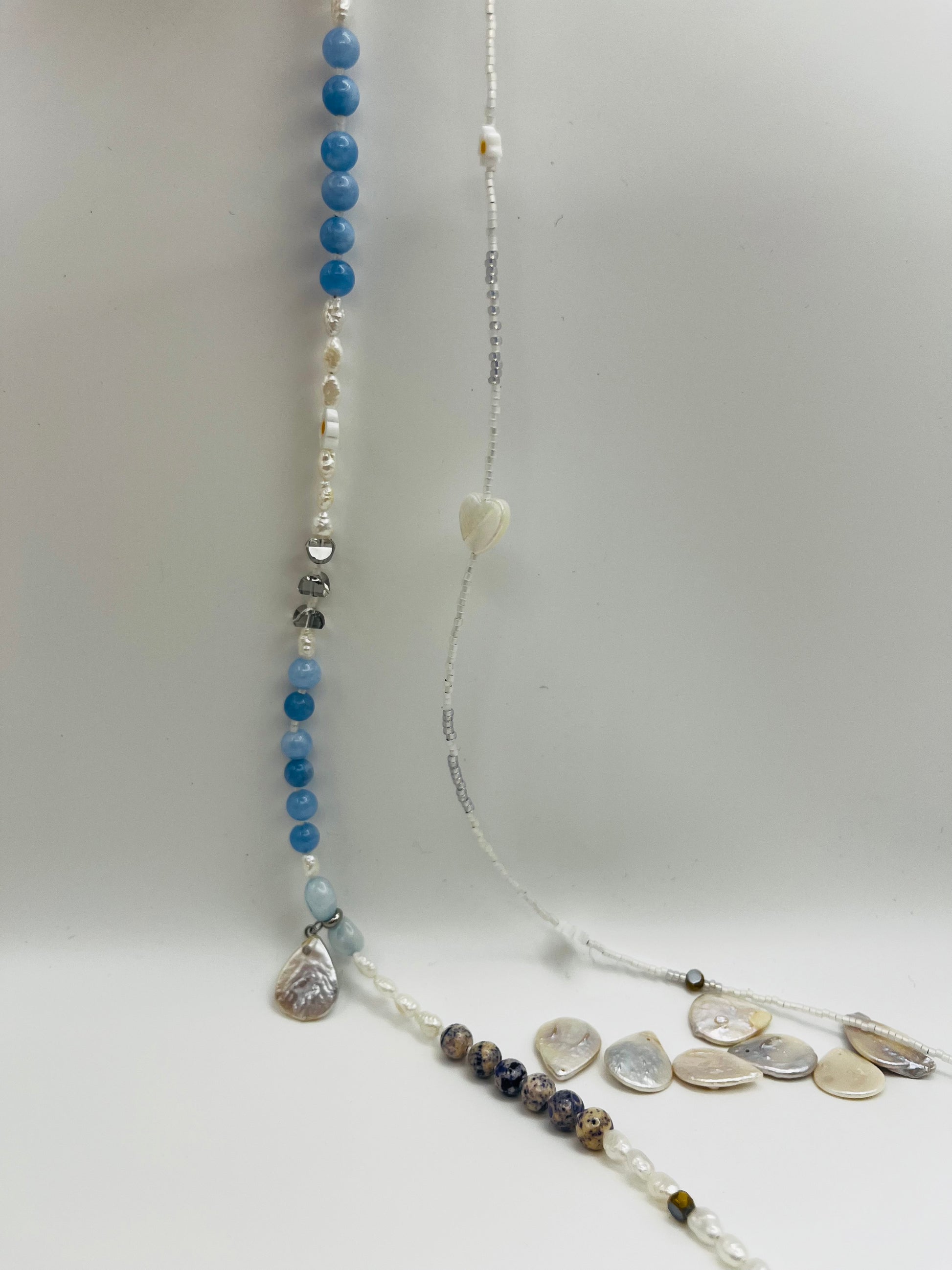 Collar Periwinkle – detalle de los dos hilos desplegados en vertical: hilo de aguamarina azul cielo facetada, perlas barrocas blancas, jaspe dálmata y cuentas plateadas con corazón colgante de cristal; e hilo de semillas de rocalla blancas con margaritas de cerámica, sobre fondo blanco, hecho a mano en Murcia