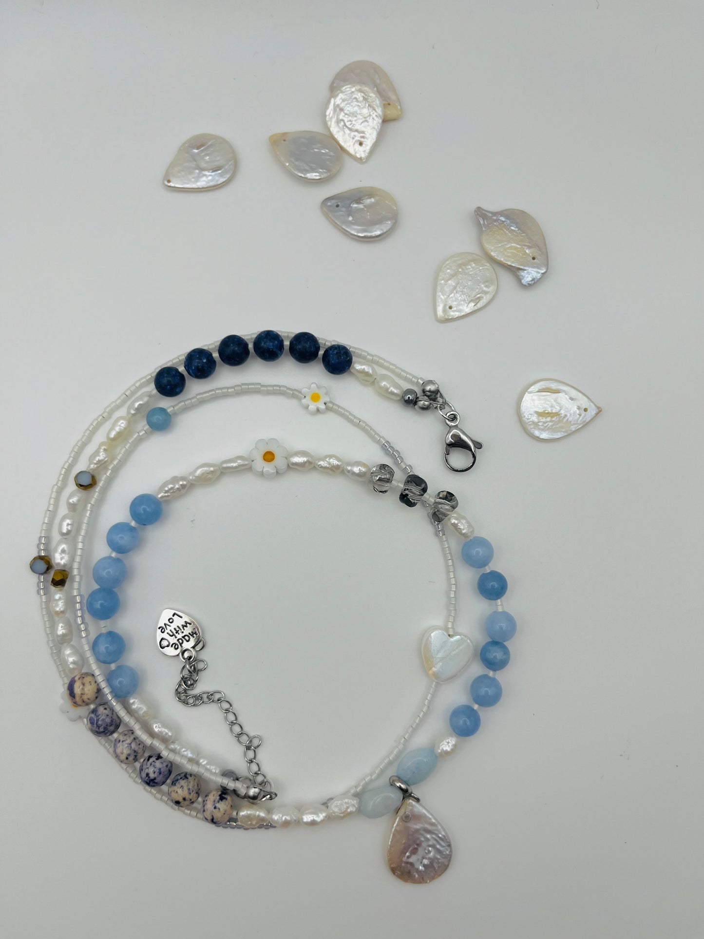 Collar Periwinkle – doble collar enrollado con semillas de rocalla blancas, margaritas de cerámica blancas, perlas barrocas blancas, aguamarina azul cielo, jaspe dálmata, cuenta negra, corazón colgante de cristal y charm "Made with Love", con perlas barrocas en forma de corazón sueltas en el fondo, hecho a mano en Murcia