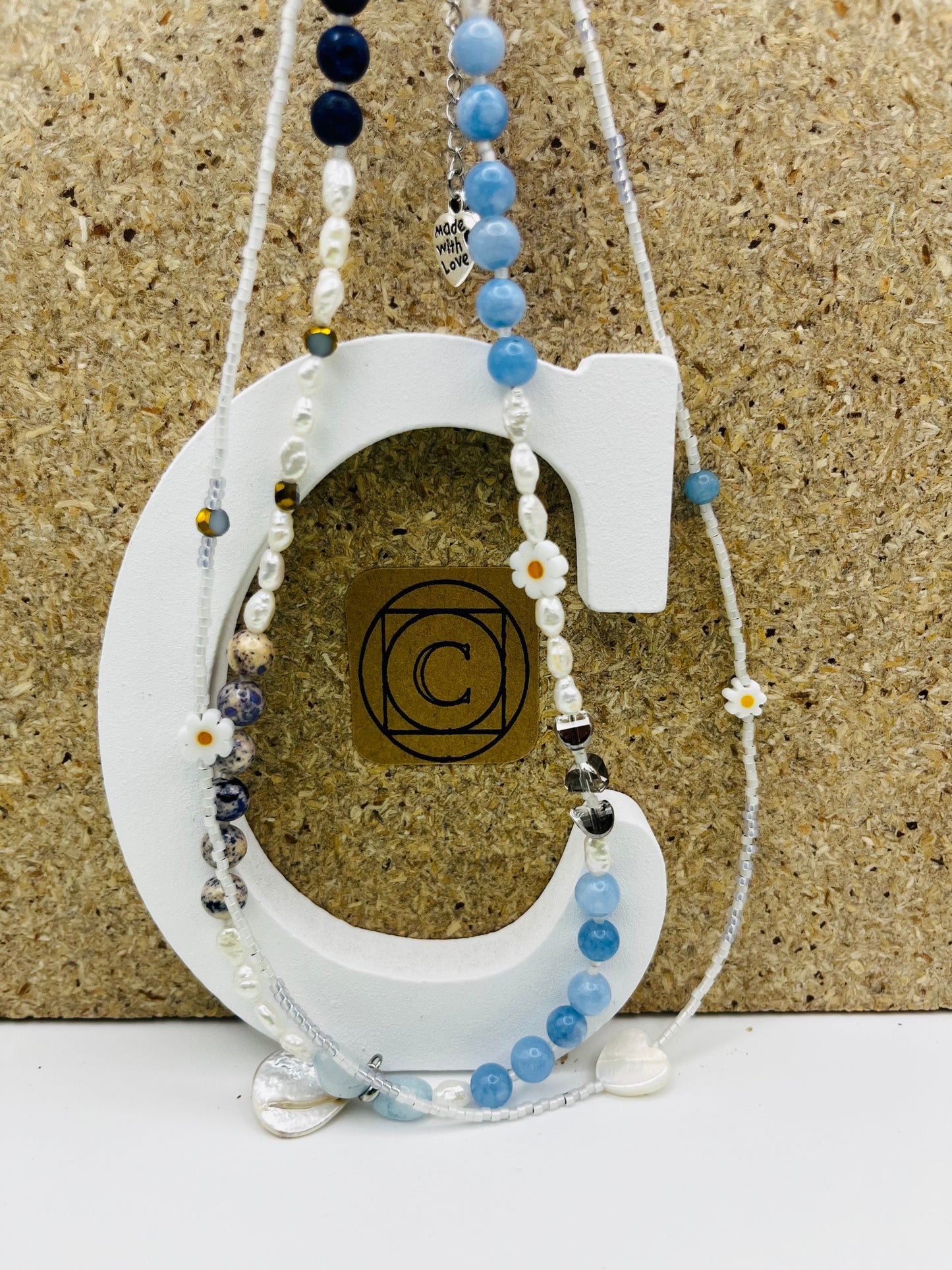 Collar Periwinkle – doble collar con semillas de rocalla blancas, margaritas de cerámica blancas con centro naranja, perlas barrocas blancas, cuentas de aguamarina azul cielo facetadas, jaspe dálmata, cuenta dorada, cuenta negra y corazón colgante de cristal transparente con charm "Made with Love", sobre letra C blanca con logo Cuadratura y fondo de corcho, hecho a mano en Murcia

